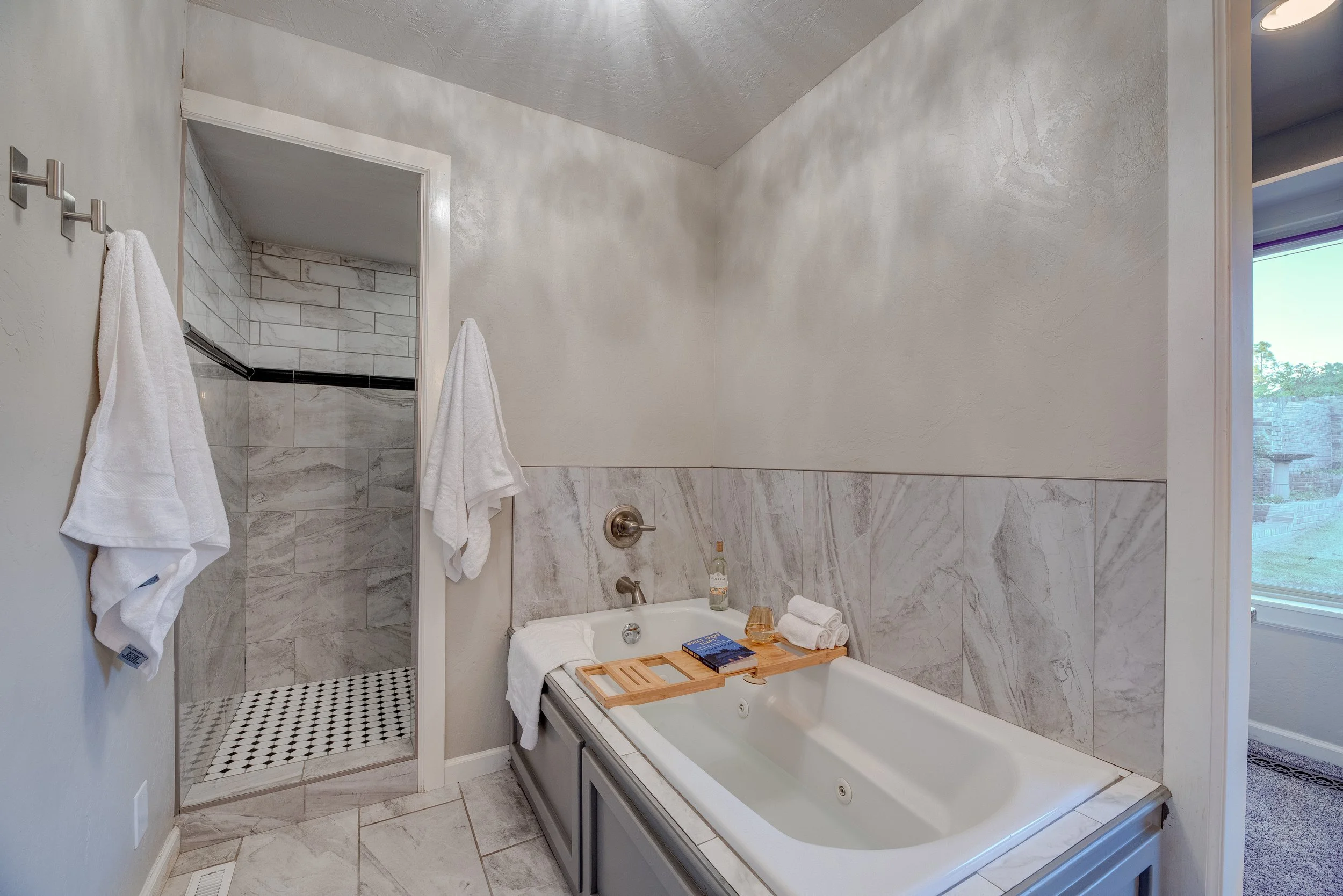 A bathroom with a bathtub, a towel rack with two towels, a tiled shower, and a window showing an outdoor view. The bathtub has a wooden tray and various toiletries.