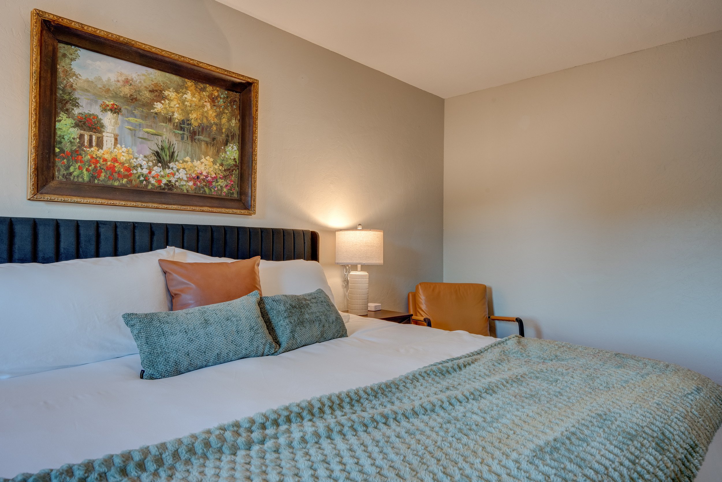 A bedroom with a large bed featuring a black headboard, pillows, and a textured blanket. There is a framed landscape painting on the wall, a bedside table with a lamp, and a tan chair.