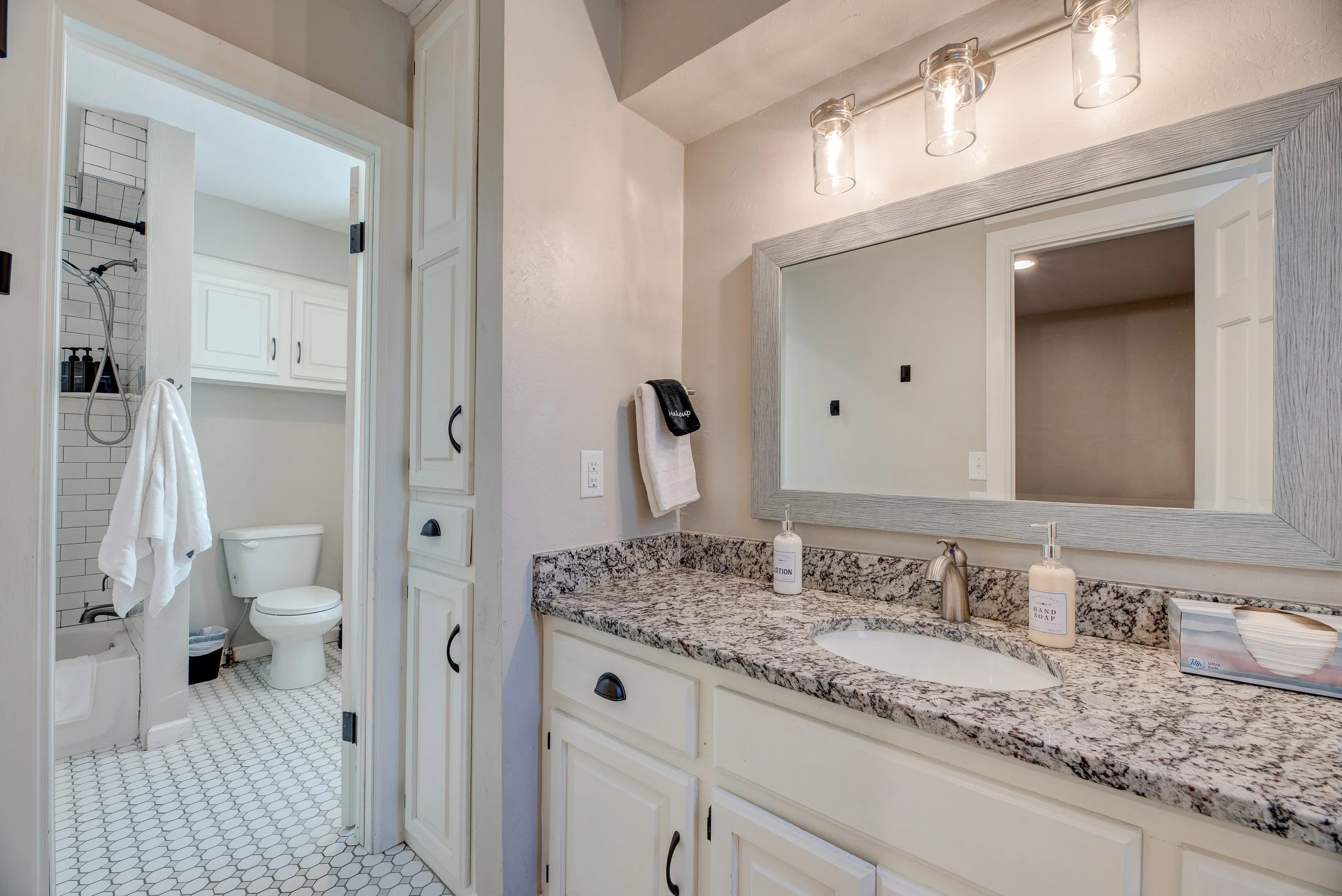 A bathroom with a granite countertop vanity, large mirror, and lighting fixture. In the background, a separate shower and toilet area are visible, with white tiles and a towel hanging.