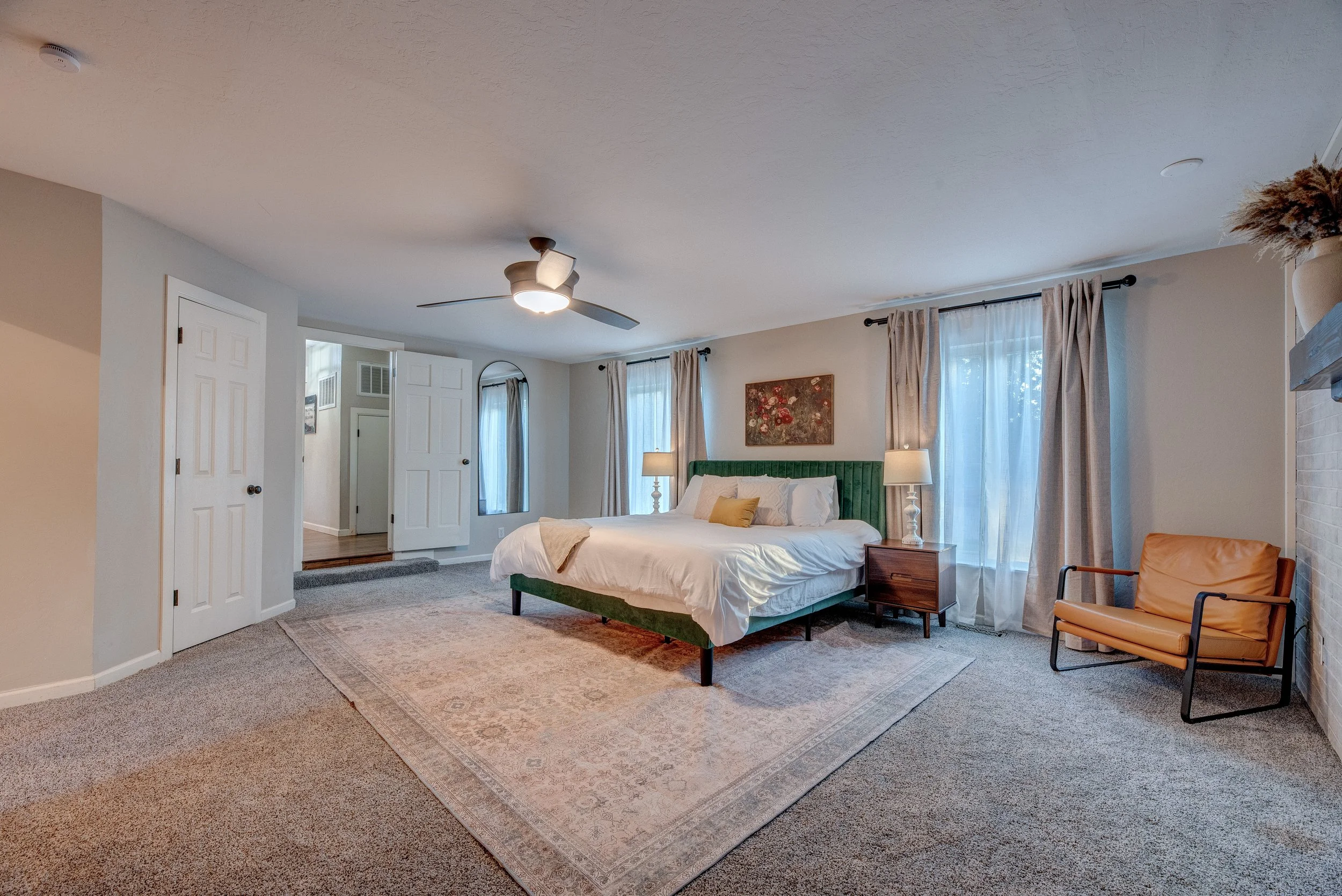 A cozy bedroom with a white bed, beige and green accents, and natural light from large windows with beige curtains. A ceiling fan is mounted on the ceiling, and there is a wooden side table with lamps and a framed painting on the wall. An orange chai