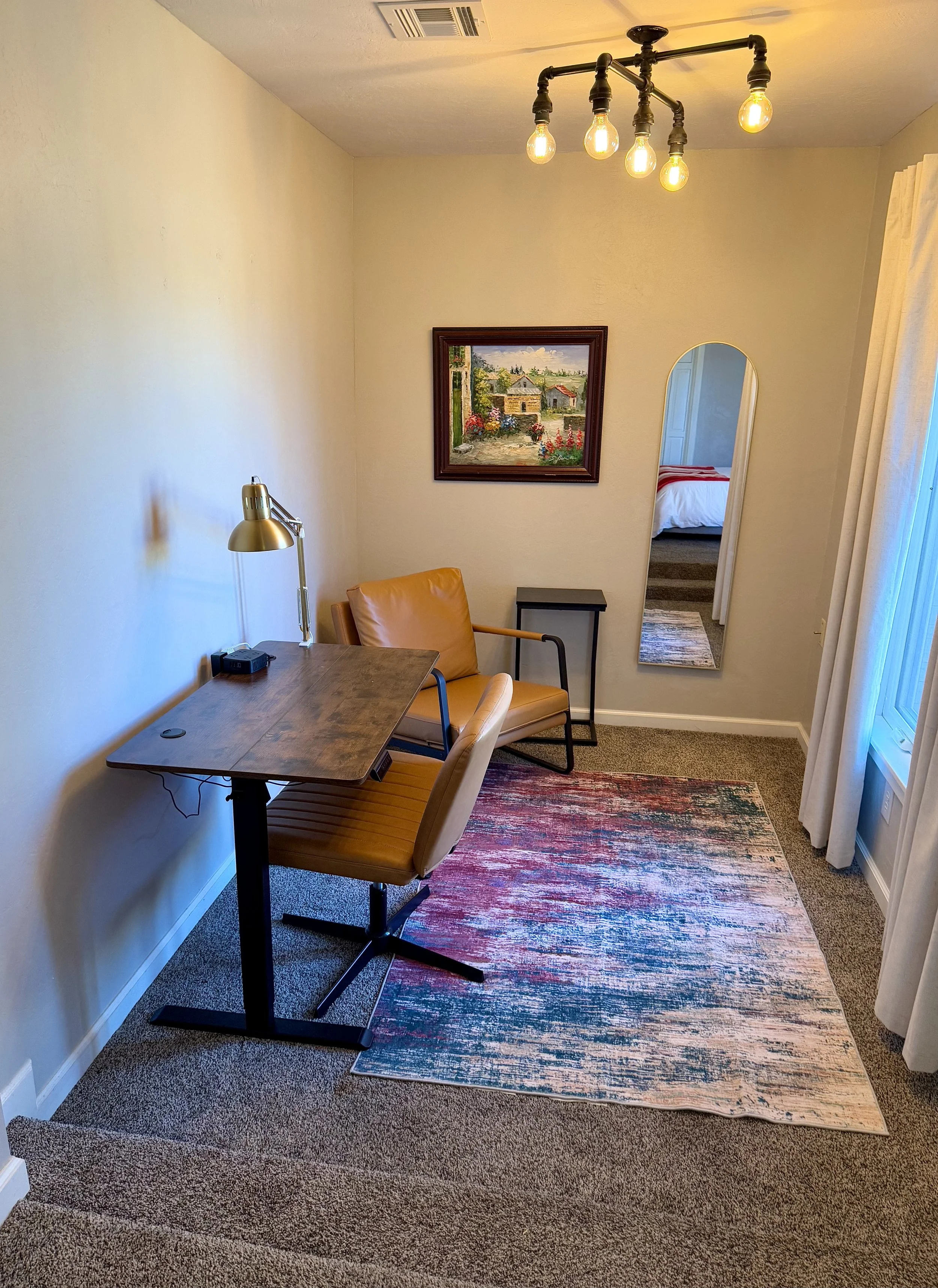 A cozy room with a wooden desk, tan chair, gold desk lamp, and small black side table. There is a large mirror on the wall and a framed painting of a landscape. Carpeted floor with colorful rug, and a half-open curtain on the window.