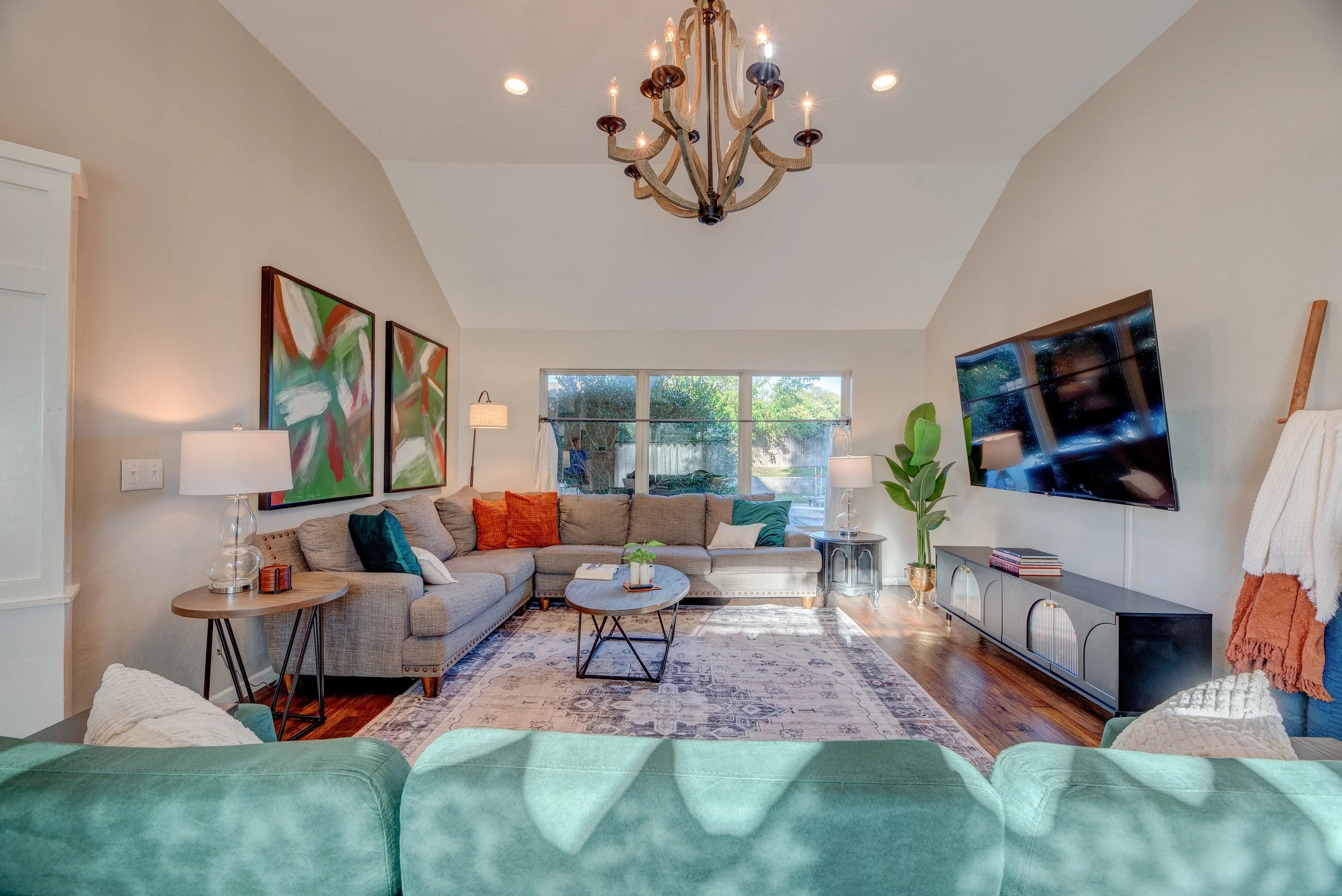 Living room with beige sectional sofa, colorful pillows, wooden coffee tables, abstract artwork, floor lamps, a large TV on the wall, a green plant, and a window showing trees outside.