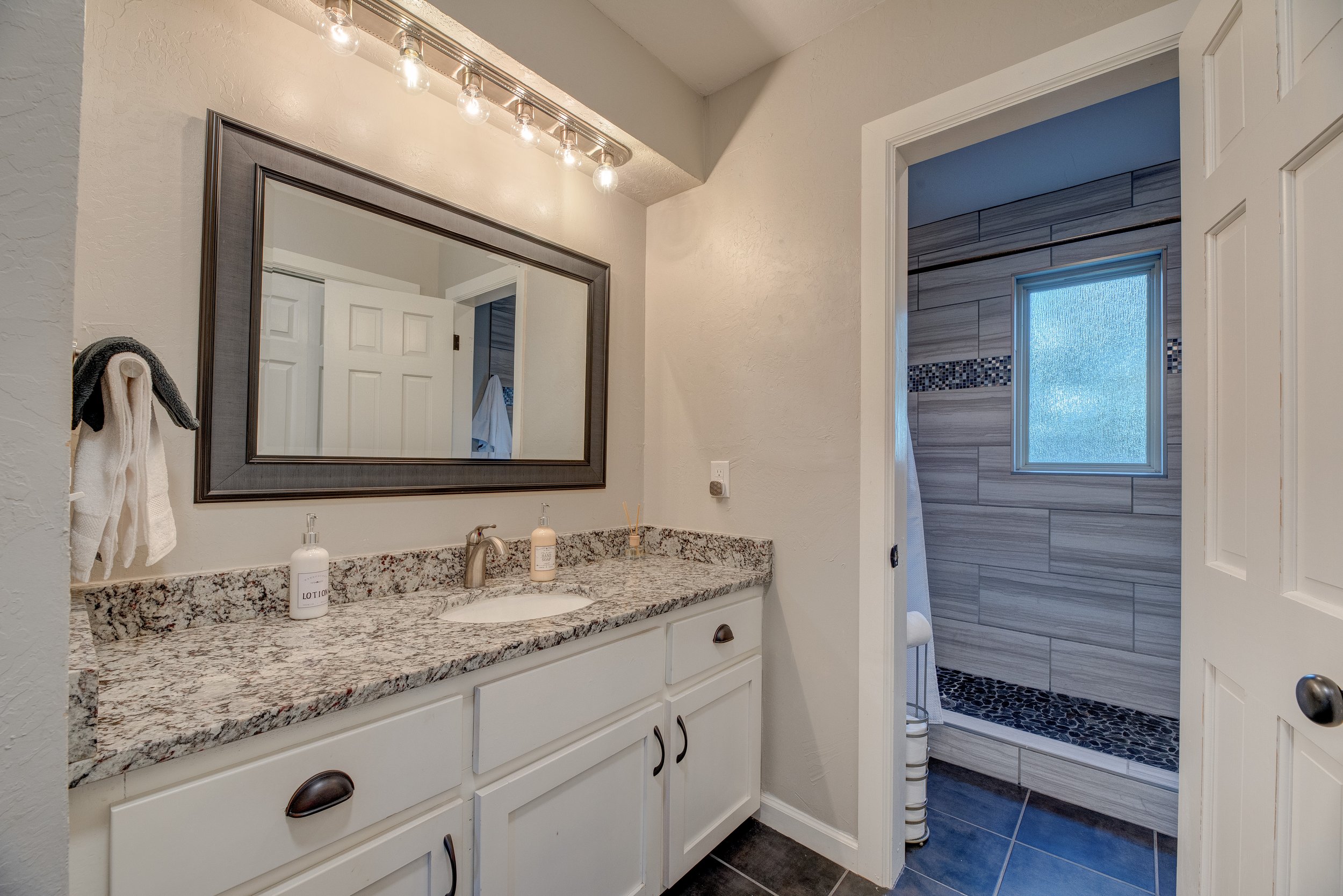 Bathroom with white vanity, granite countertop, large mirror, and a shower with textured glass window and gray tile walls.