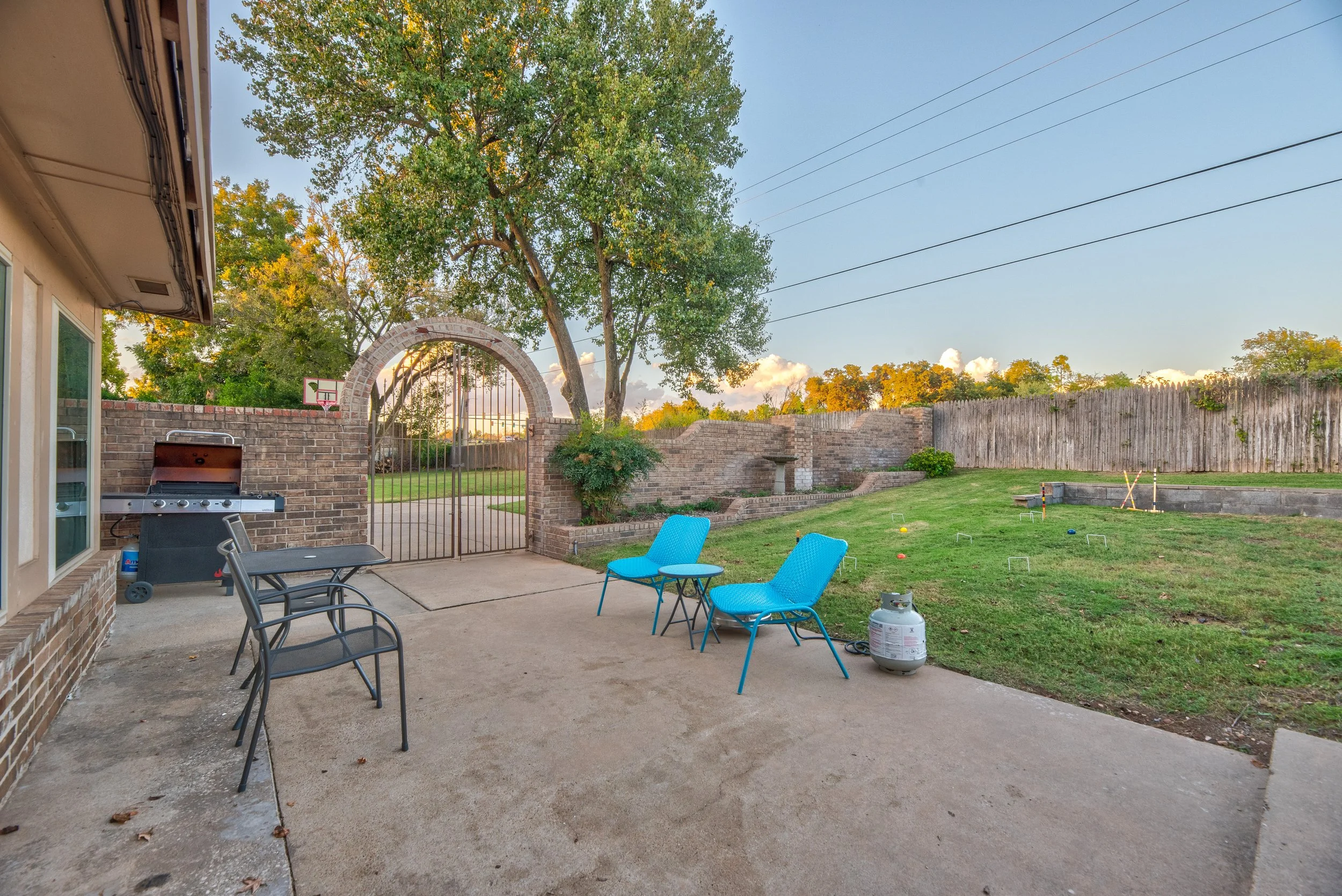Backyard patio with outdoor chairs, propane tank, and barbecue grill, enclosed by brick wall and wooden fencing, with a lawn and mature trees in the background under a evening sky.