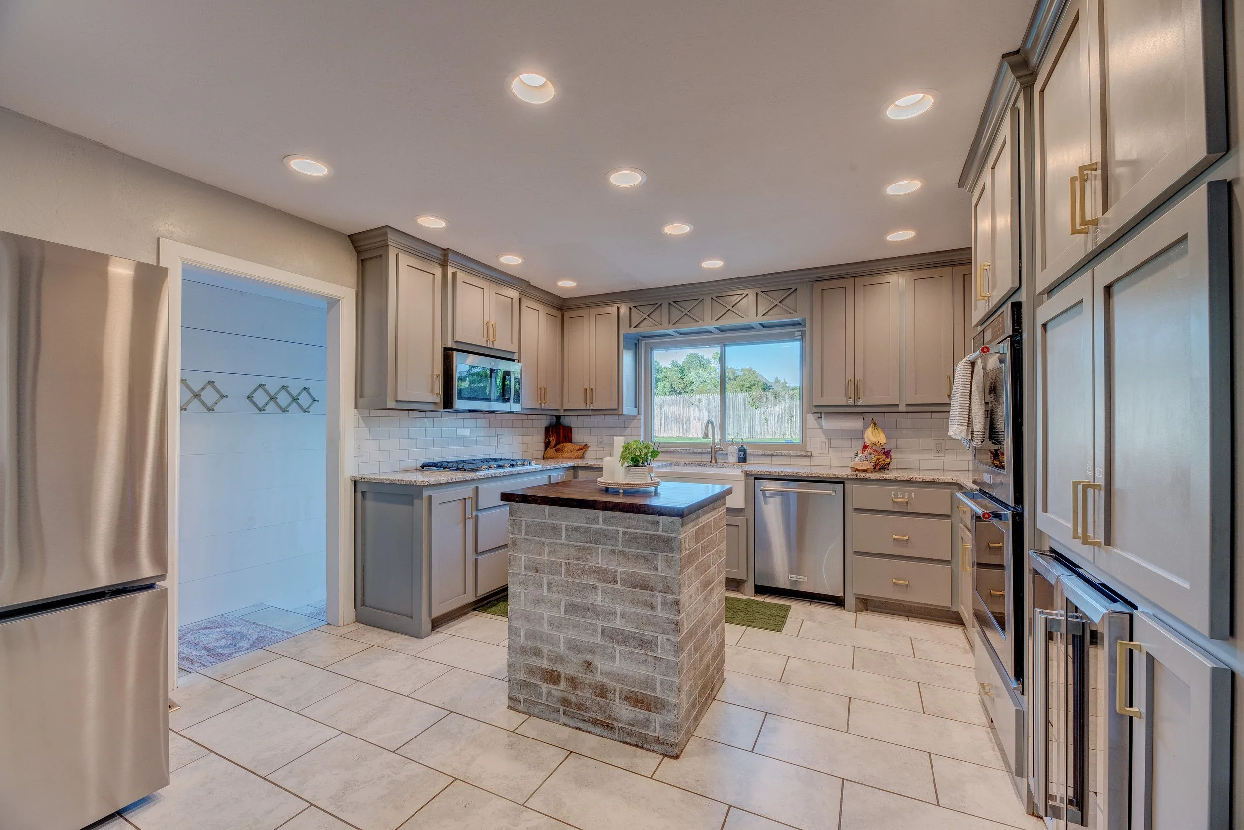 A modern kitchen with light gray cabinets, a brick island, stainless steel appliances, a window above the sink, and recessed ceiling lights.