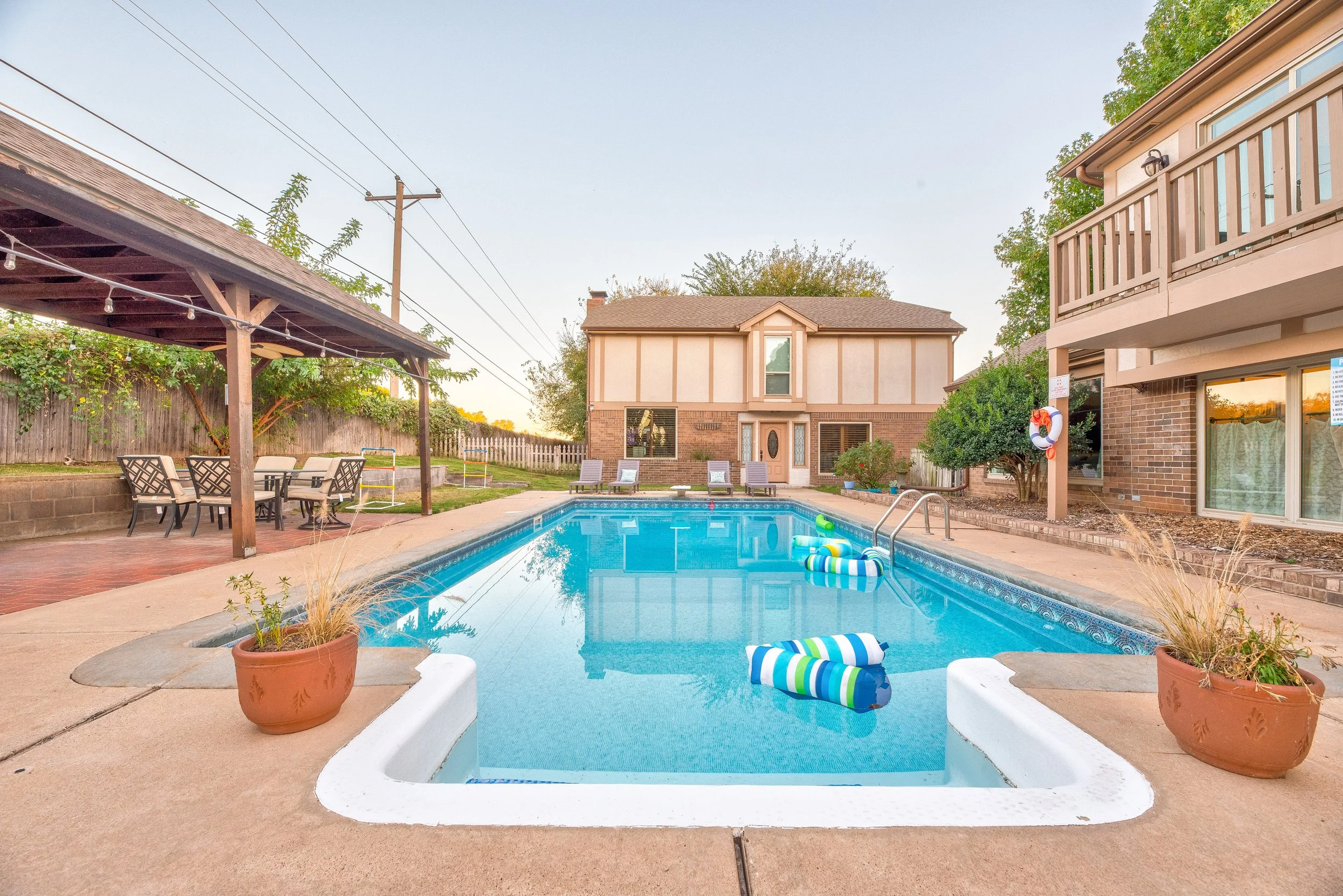 Residential backyard with a swimming pool, patio furniture, potted plants, and surrounding trees and houses during sunset.