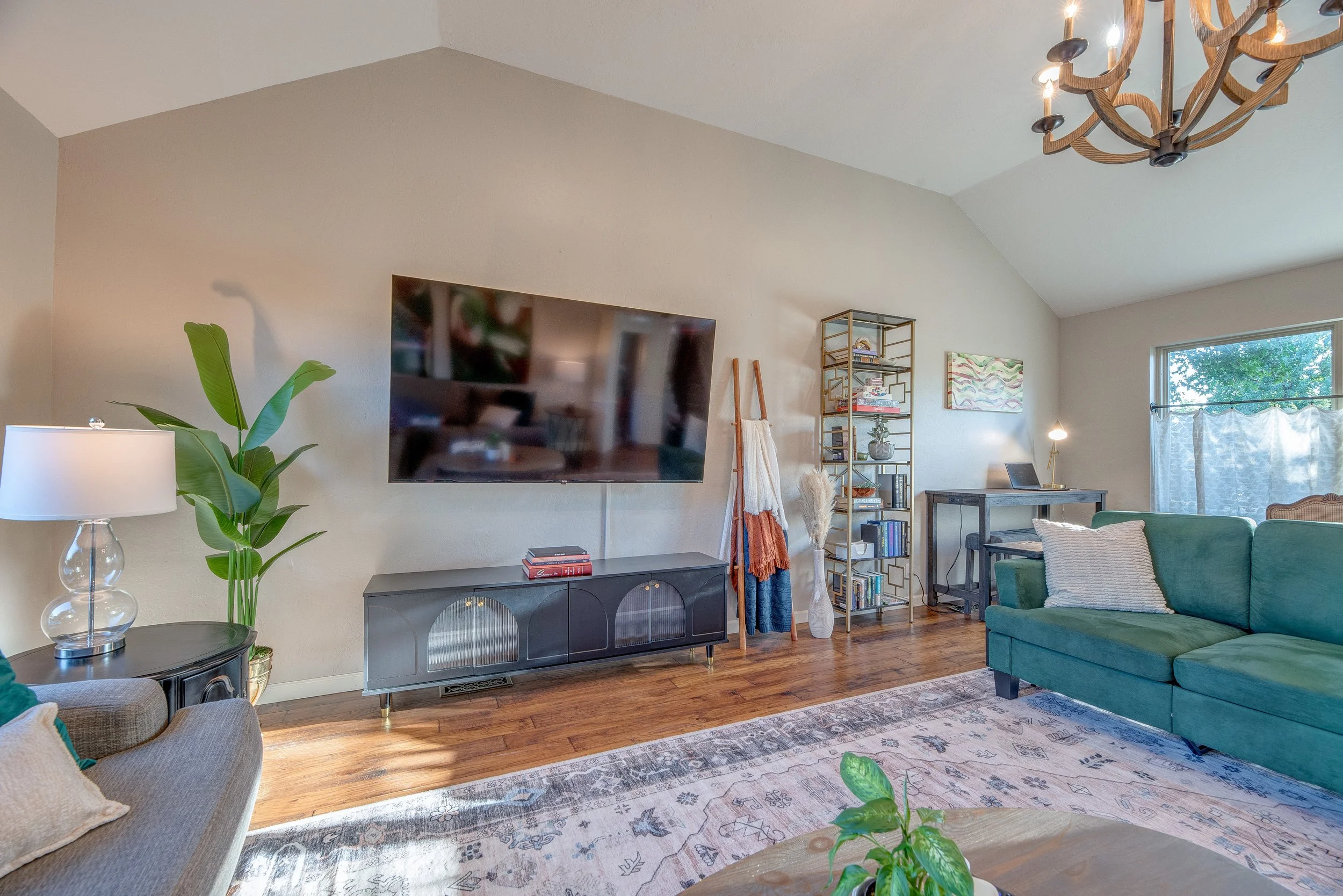 Living room with a large flat-screen TV on a stand, a green sofa with pillows, a patterned rug, a side table with a lamp, a bookshelf filled with books and decorations, and a window with curtains.