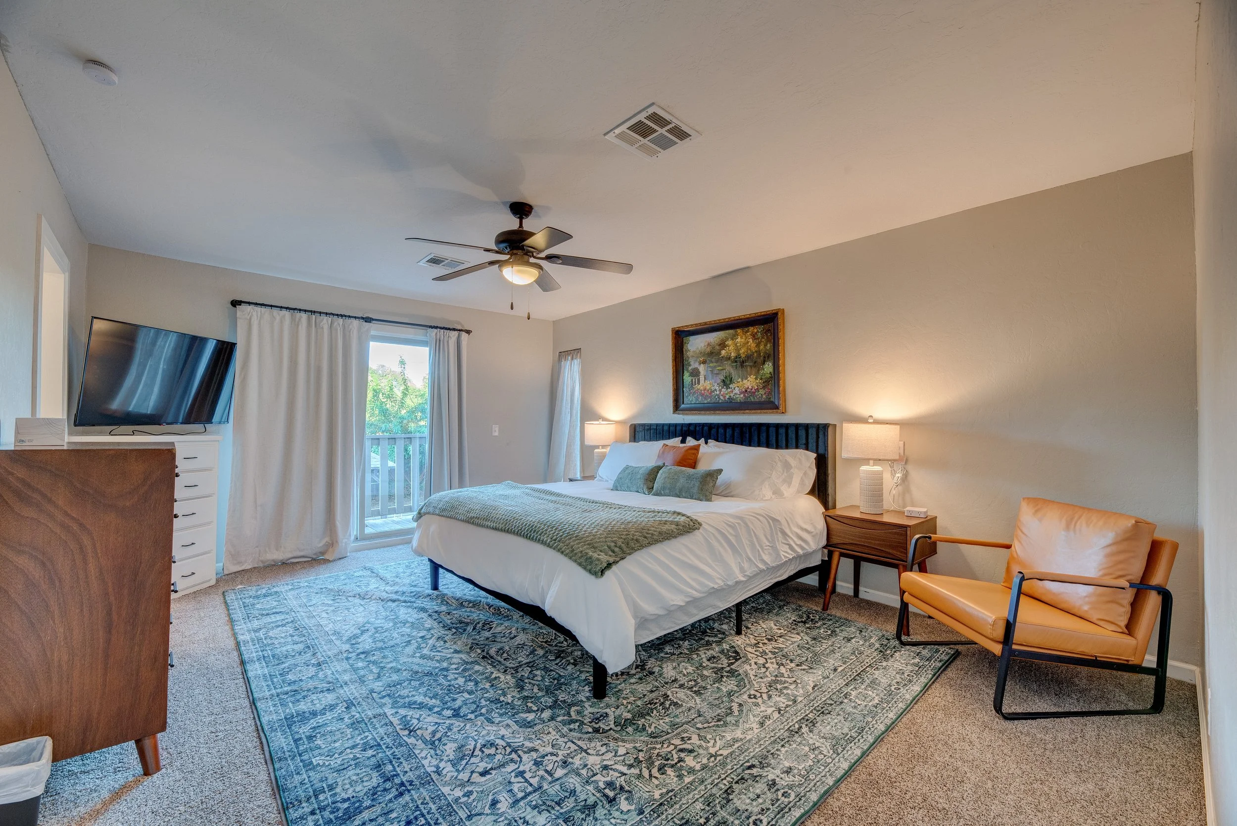 A hotel room with a large bed, a painting above the bed, a window with white curtains, a ceiling fan, a TV mounted on the wall, a wooden dresser, and an orange chair.