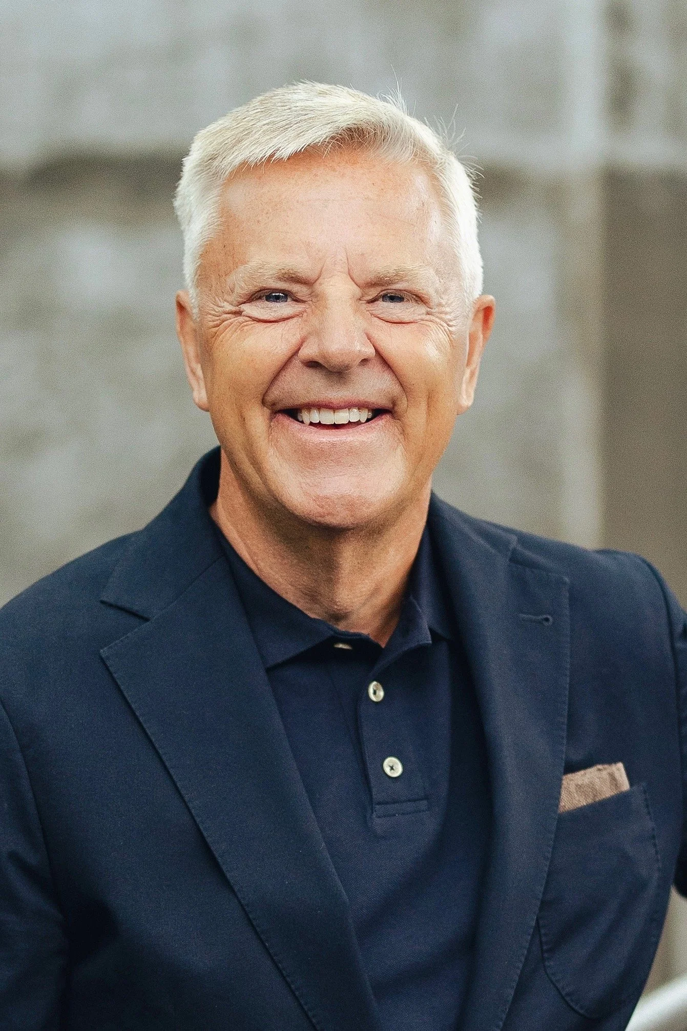 En äldre man med vitt hår, mörkblå kostym och leende.
