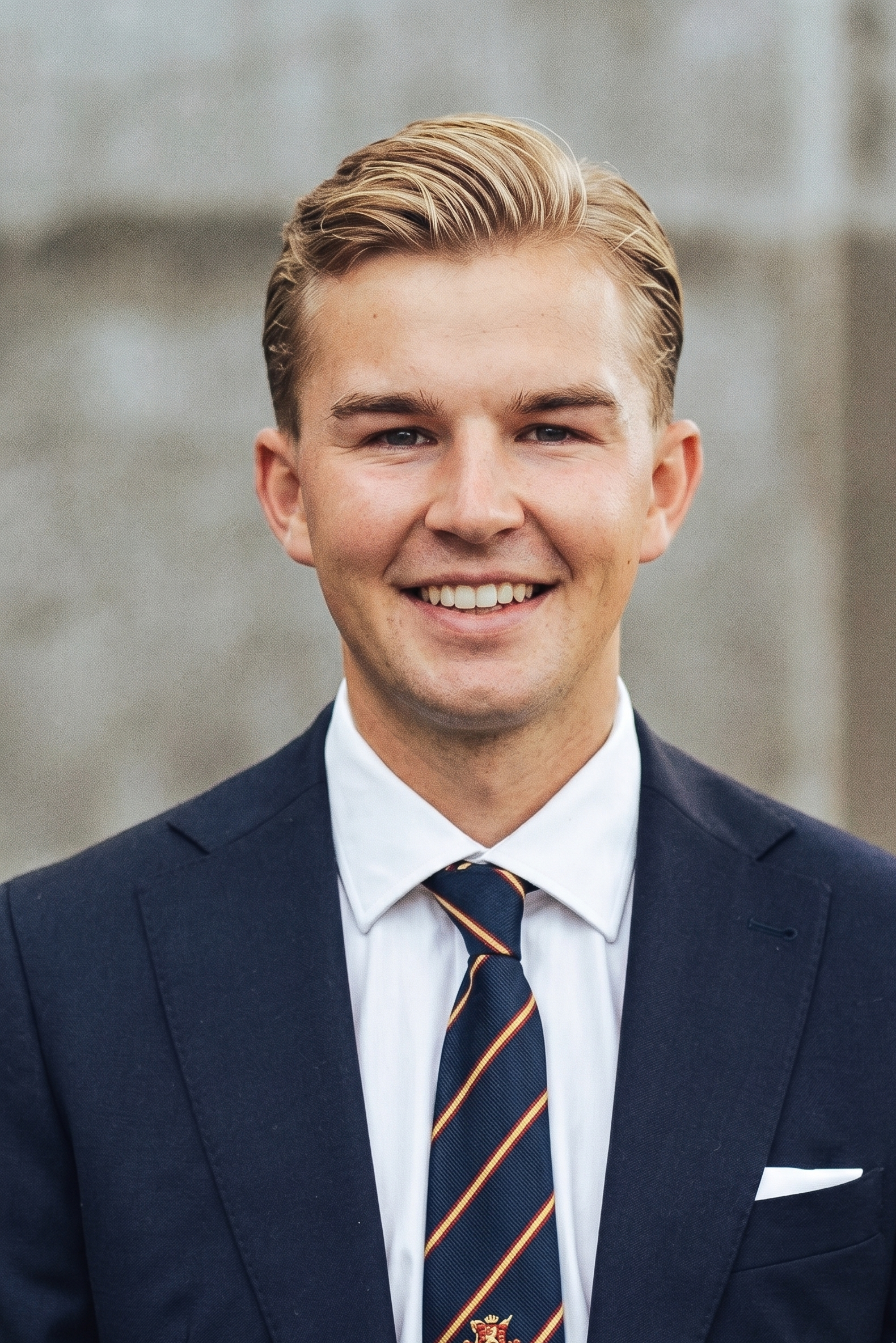 En ung man i kostym, vit skjorta och randig silkeslips, leende, med kort blont hår. Bakgrund av grå sten.