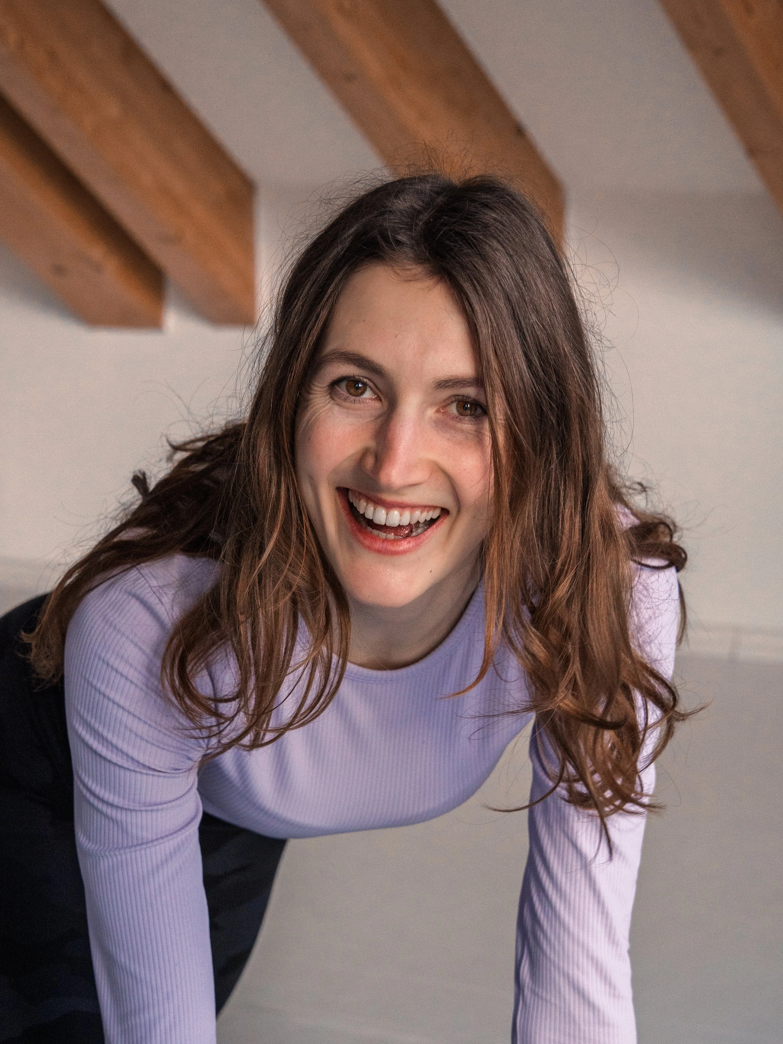 Junge Frau mit braunen, gewellten Haaren lächelt freundlich nach oben gerichtet in einem freundlichen Yoga-Raum mit Holzdeckenbalken.