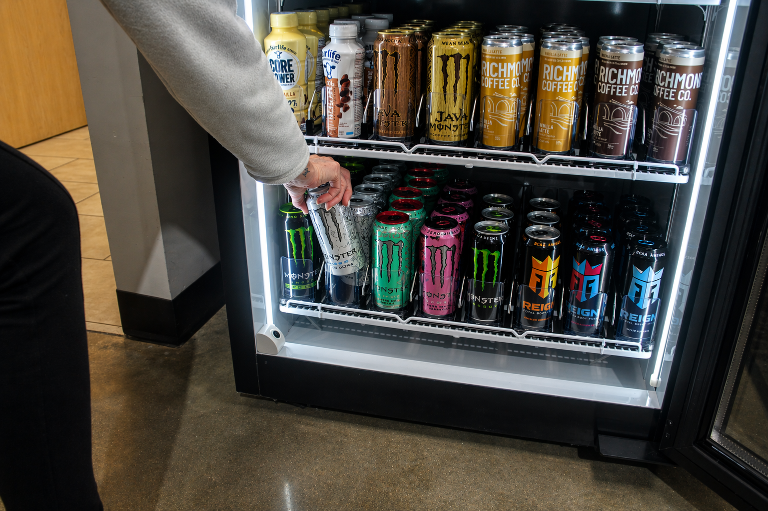 A person selecting a can of Monster energy drink from a vending machine filled with various energy drinks.