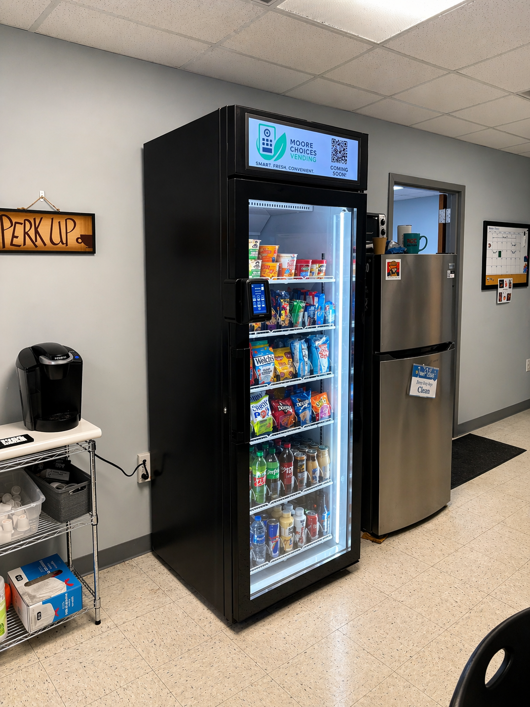 A vending machine filled with snacks and beverages, next to a stainless steel refrigerator and a coffee machine on a small table in a break room.
