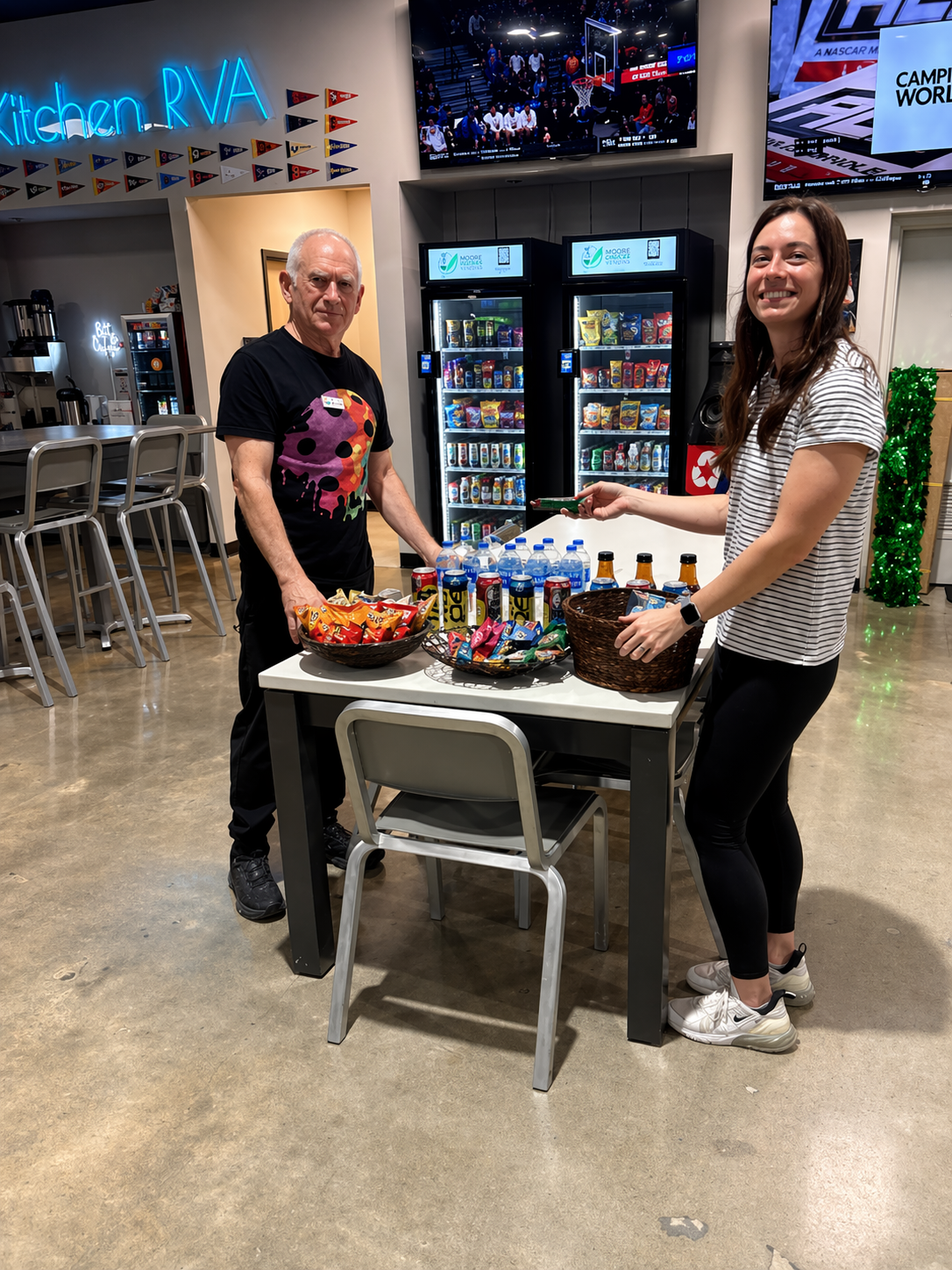 A man and a woman at a table with snacks and drinks in a lounge area, with vending machines and a TV screen in the background.