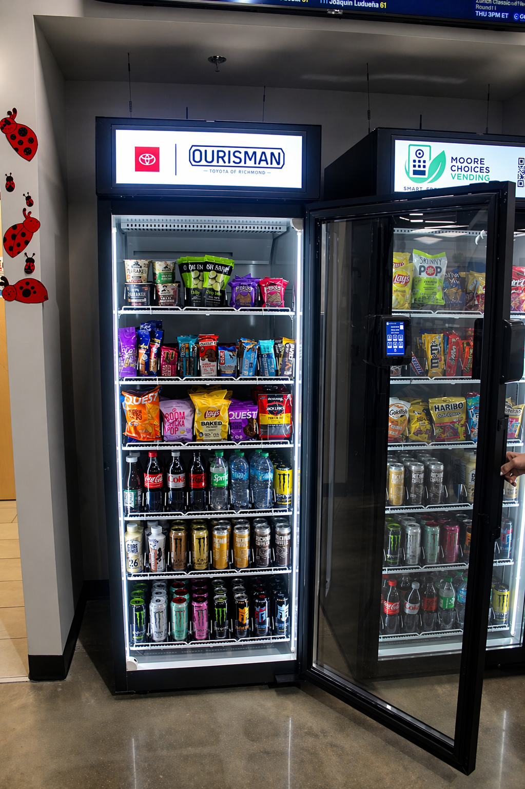 A vending machine filled with snacks and beverages, located in an indoor space. The machine has a sign on top that reads 'OURISMAN Toyota of Richmond.' Snacks such as chips and candy are on the shelves, and soda cans and bottled drinks are on the lower shelves.