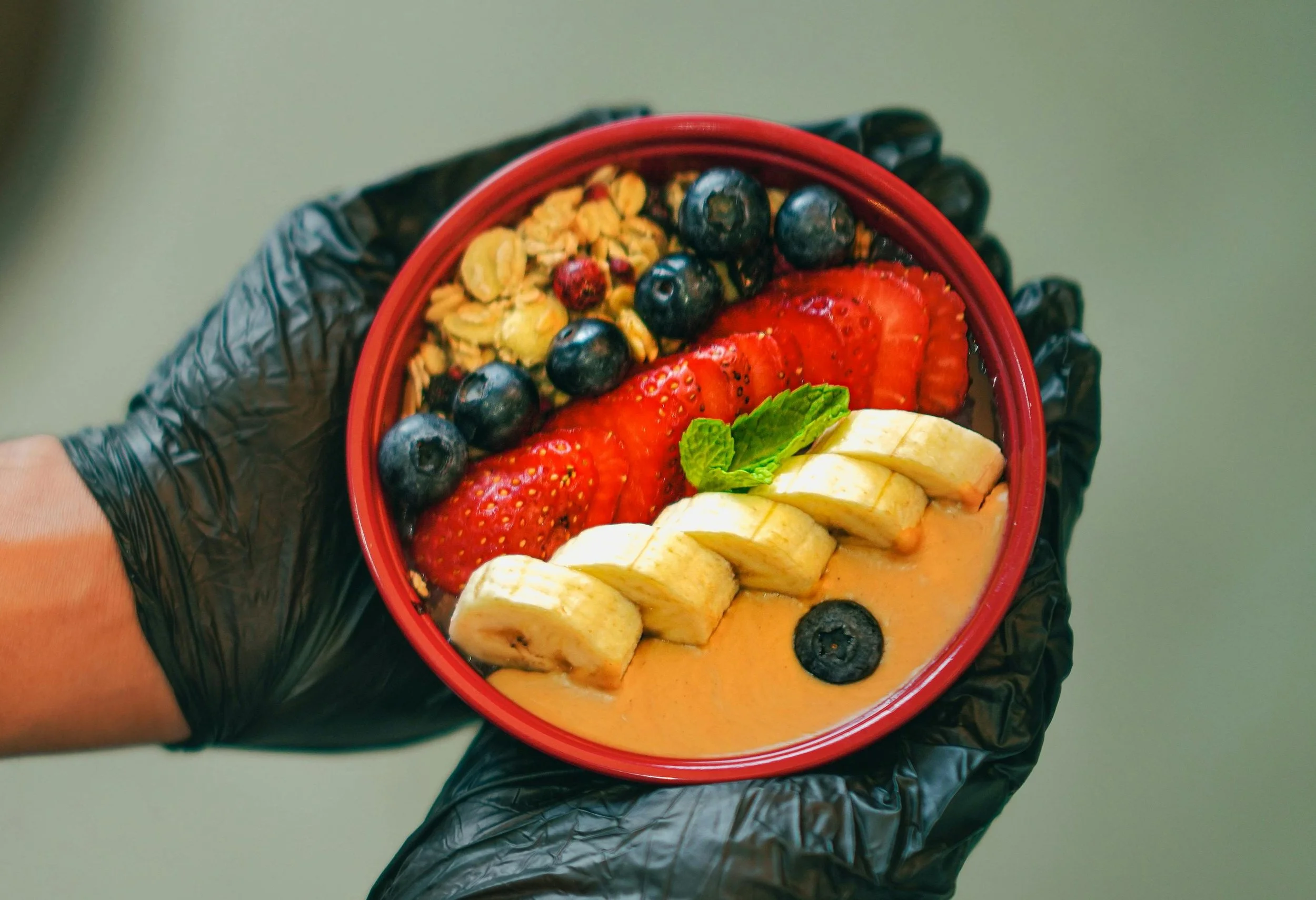 Eine handhaltende Schüssel mit Schichten aus Früchten, darunter Erdbeeren, Bananen, Heidelbeeren, Granola und Minze, in einem schwarzen Handschuh.