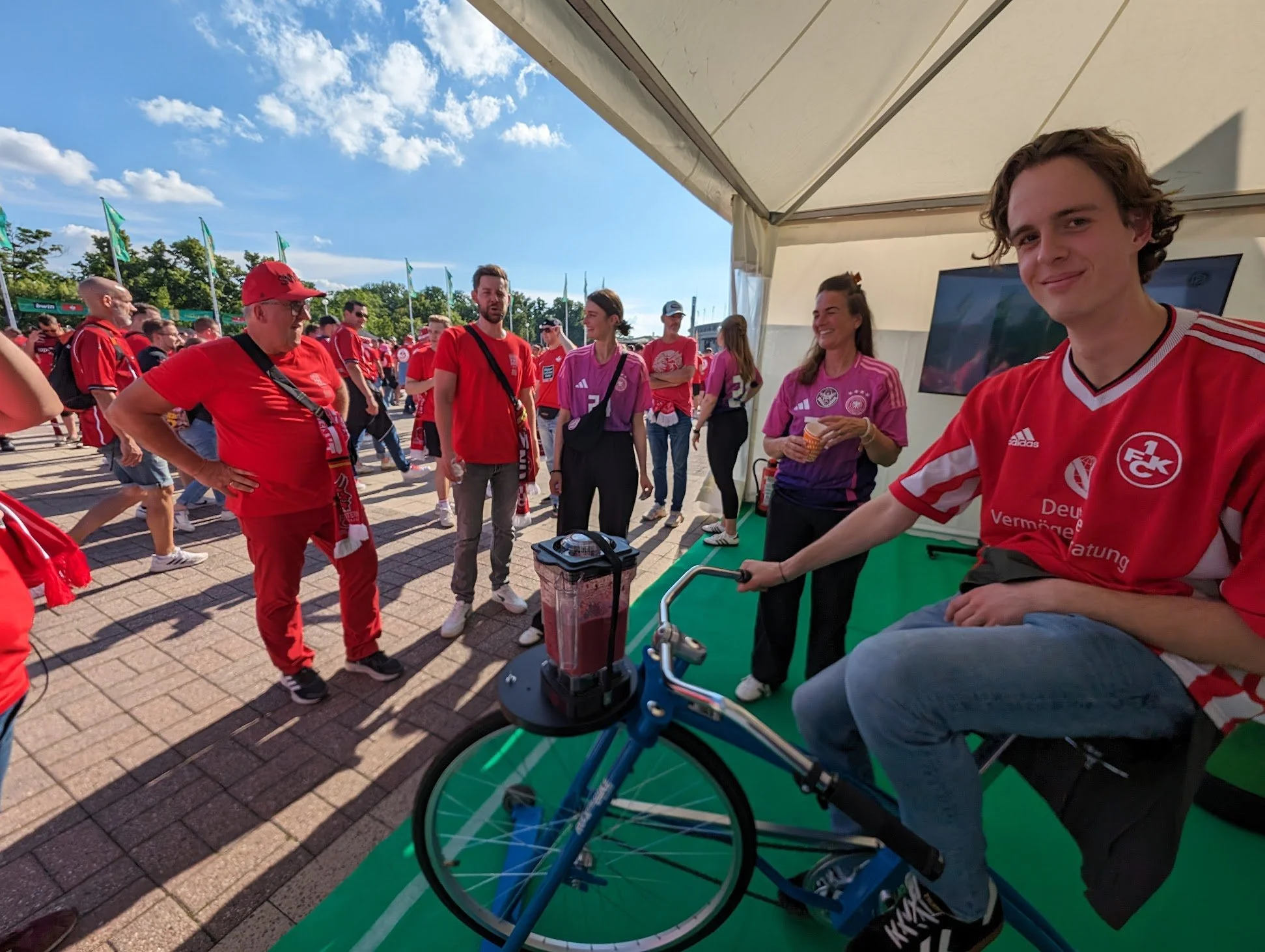 Ein junger Mann in einem roten Fußballtrikot sitzt auf einem stationären Fahrrad, während eine Gruppe von Menschen im Hintergrund bei einem Fußballereignis versammelt ist. Es ist sonnig, und die Szene findet unter einem Zelt statt.