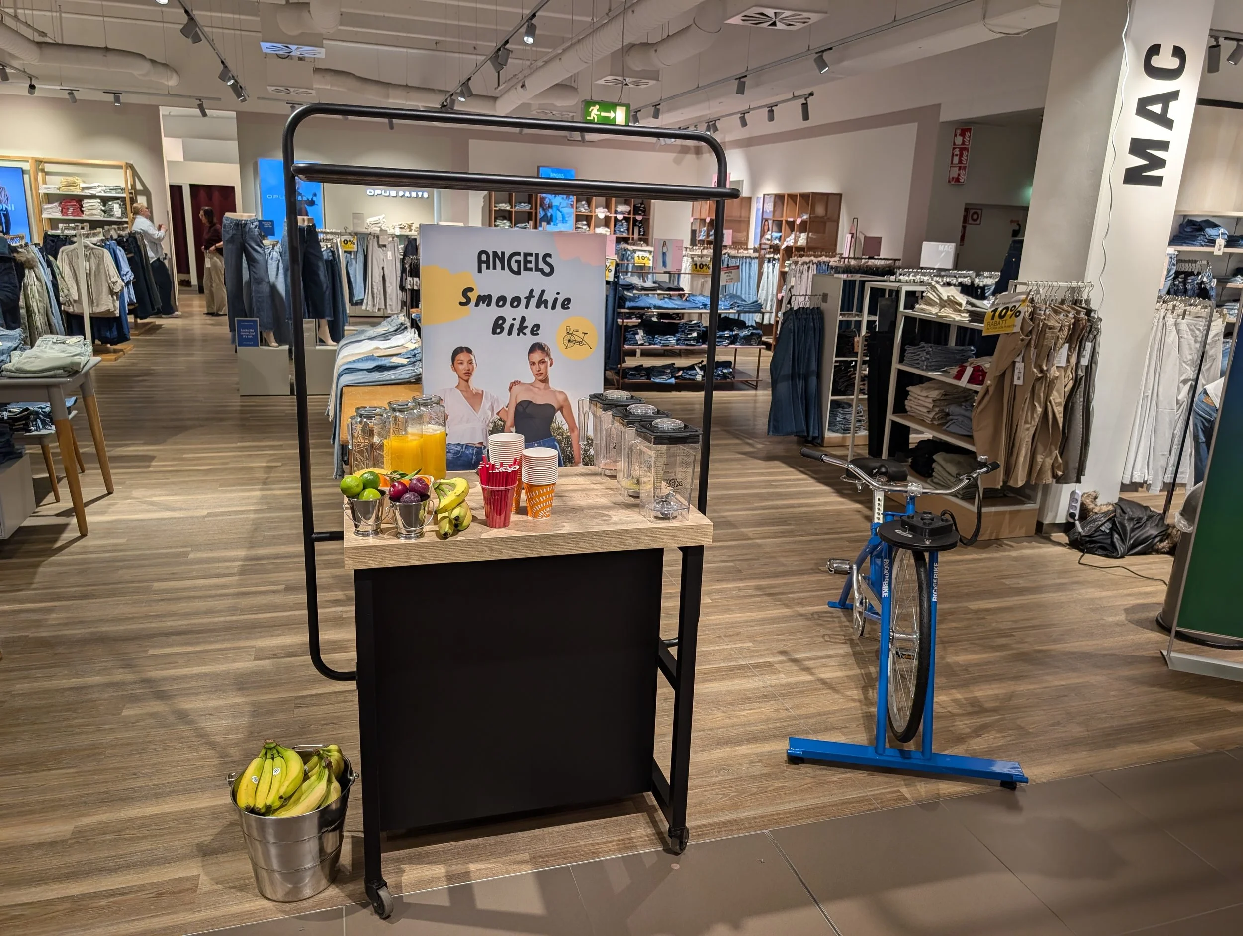 Ein Verkaufsstand in einem Bekleidungsgeschäft mit einer Plakatbeschreibung für eine Smoothie-Bike-Aktion, frisches Obst, Getränke und Kaffeepappteller auf einer Theke, Fahrräder und Bekleidung im Hintergrund.