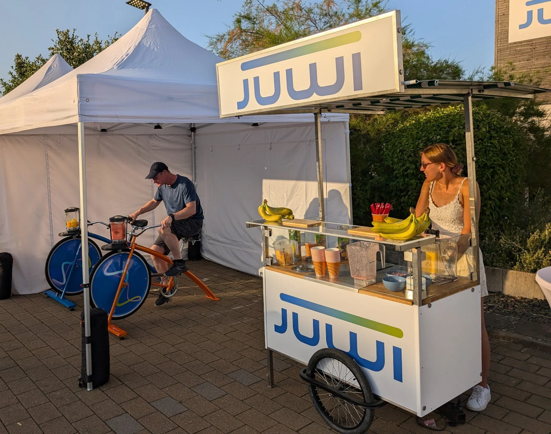 Ein Stand mit Bananen und Pappbechern, eine Frau steht hinter dem Stand, ein Mann sitzt auf einem festen Fahrrad neben einem weißen Zelt.