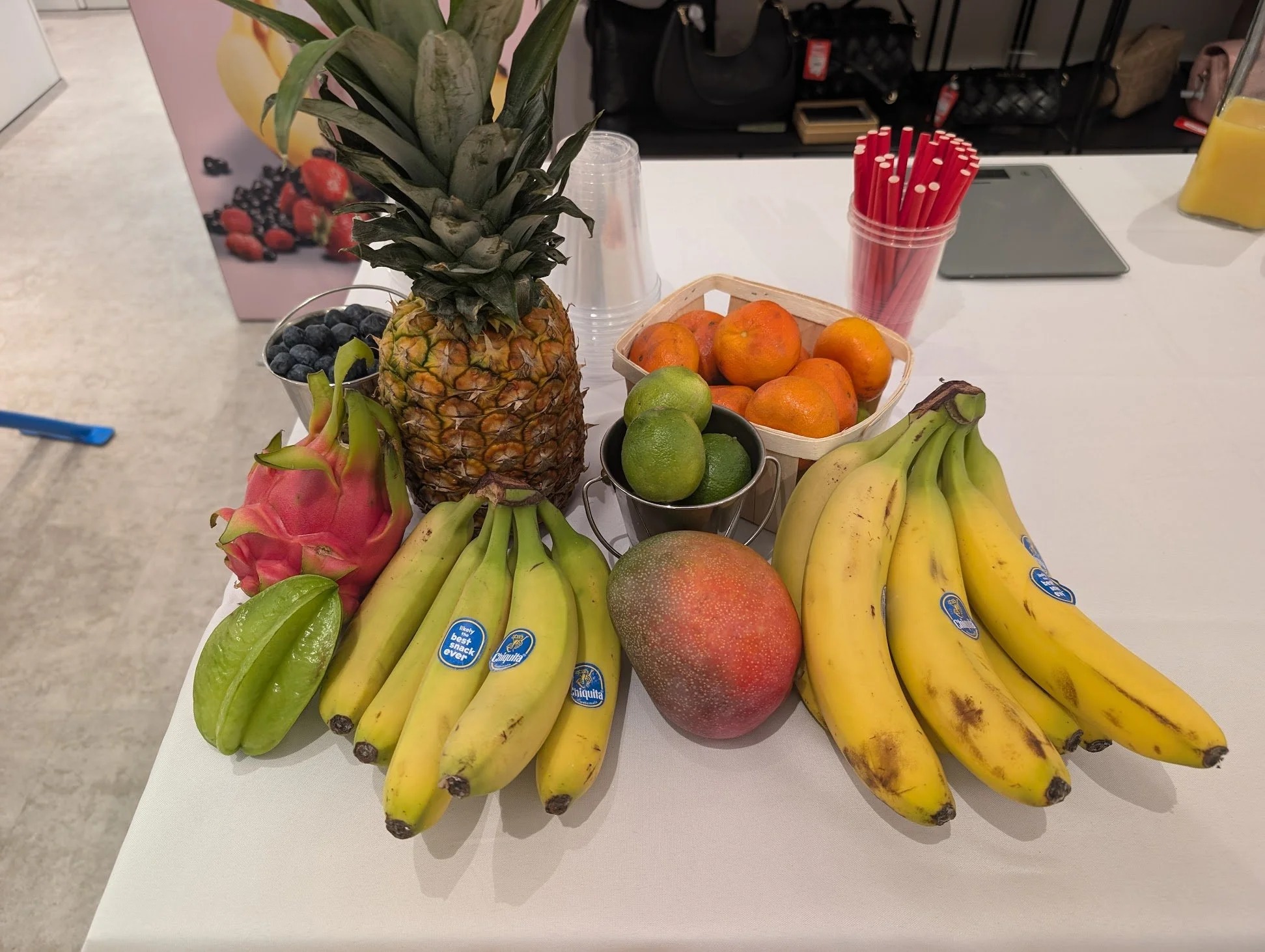 Verschiedene Früchte auf einem weißen Tisch, darunter Ananas, Bananen, Mango, Limetten, Orangen, Drachenfrucht, Heidelbeeren, und andere Früchte, mit roten Trinkhalmen im Hintergrund.