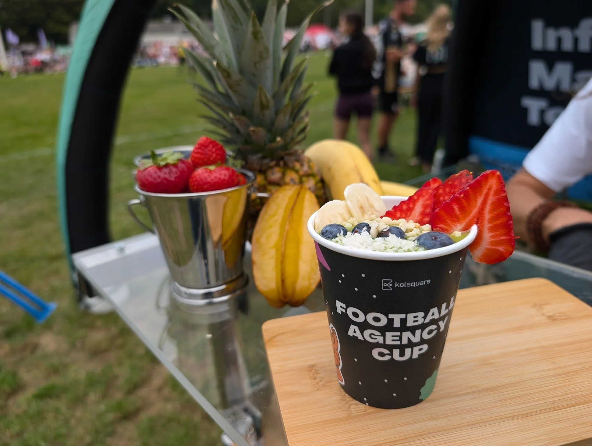 Fruchtbecher mit Erdbeeren, Banane, Heidelbeeren und Kokosraspeln, daneben eine Metalltasse mit Erdbeeren sowie eine Ananas und weitere Früchte im Hintergrund auf einem Tisch bei einem Outdoor-Event.