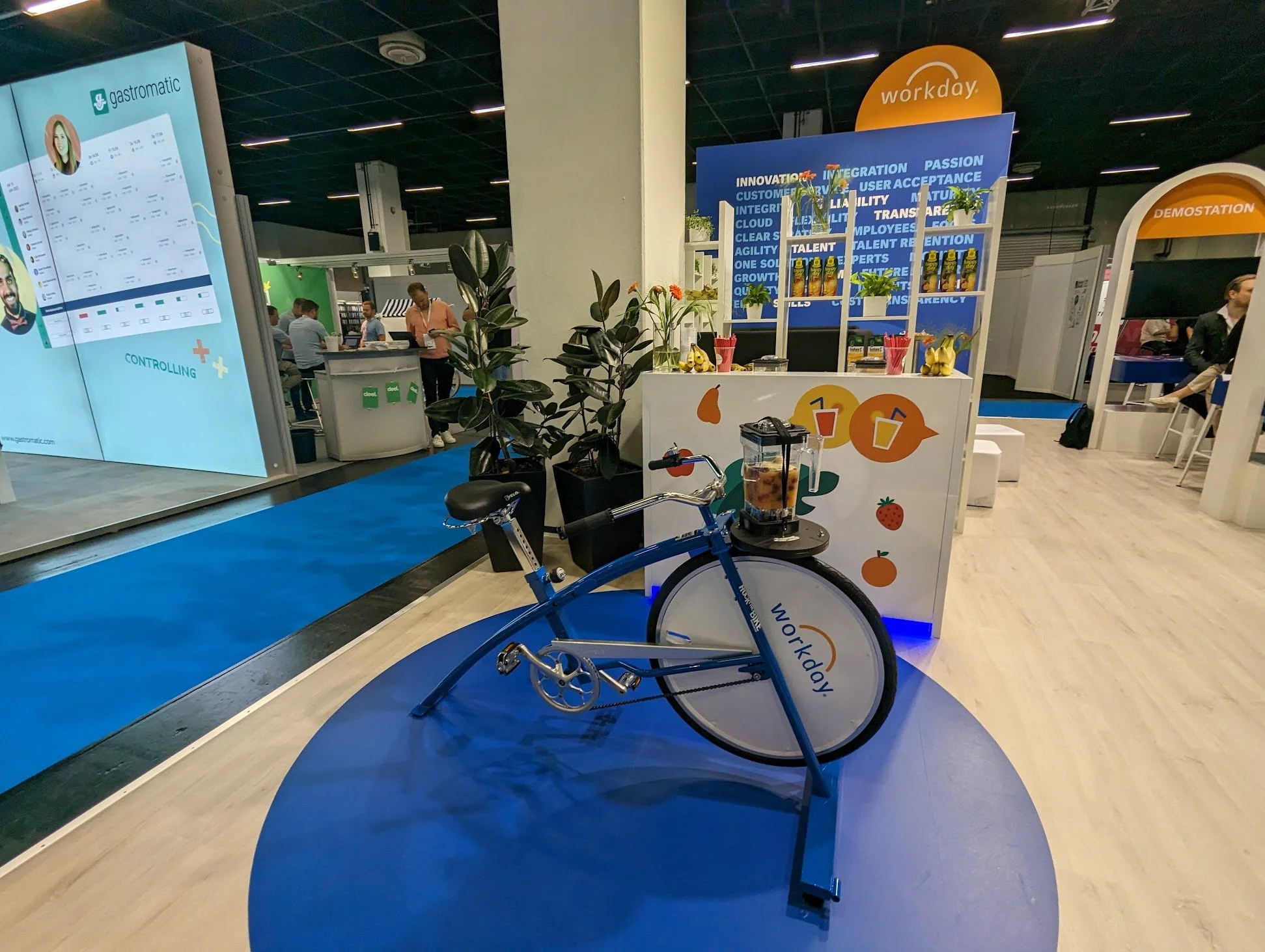 Ein blauer Indoor-Fahrradtrainer mit einem Getränkehalter auf einem Plattform, im Hintergrund ein Messestand mit Pflanzen und mehreren Personen bei einer Messe.