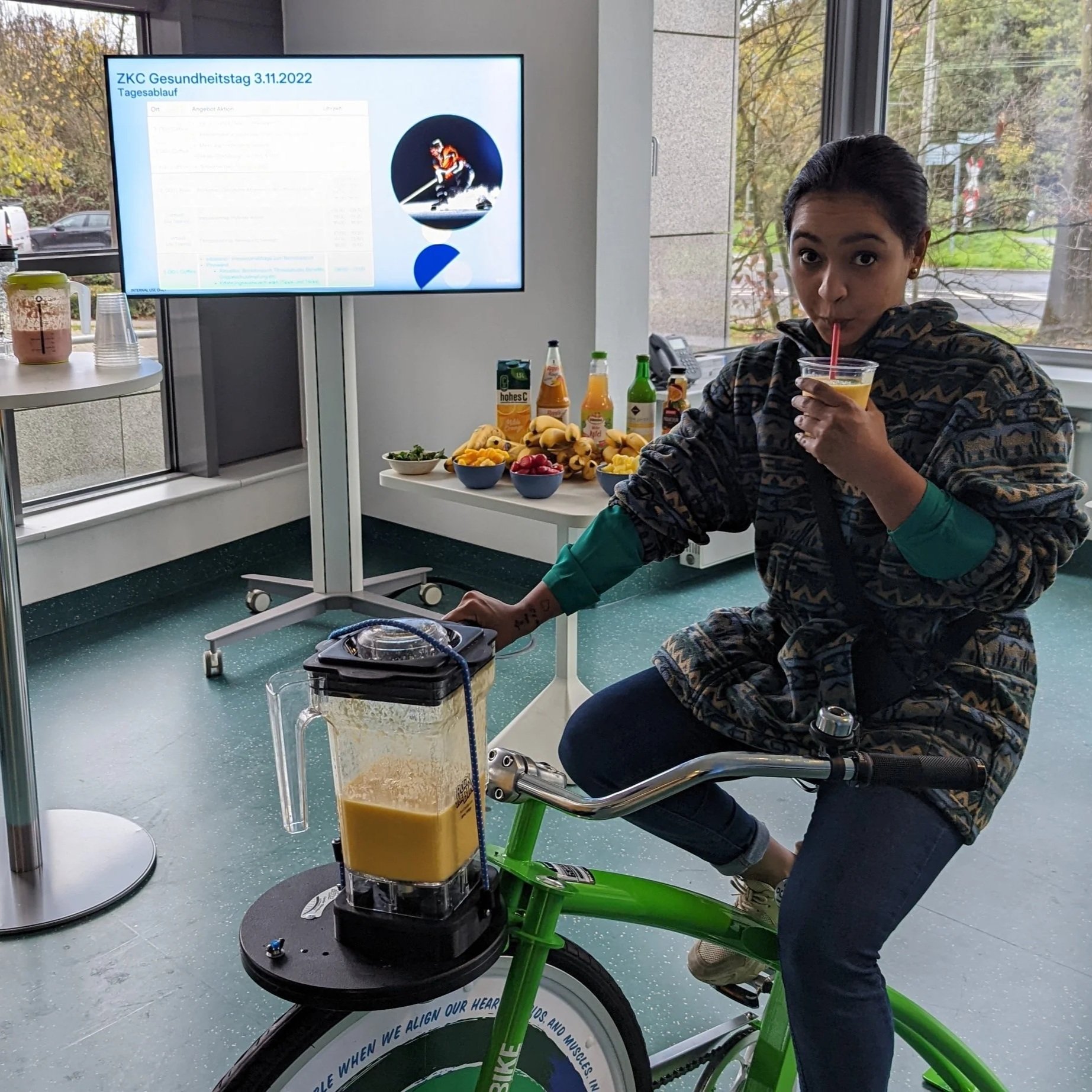 Junge Frau auf einem grünen Fahrrad im Raum neben einem großen Bildschirm, Getränke auf einem Tisch im Hintergrund, eine Flüssigkeit in einem Mixer, die sie trinkt.