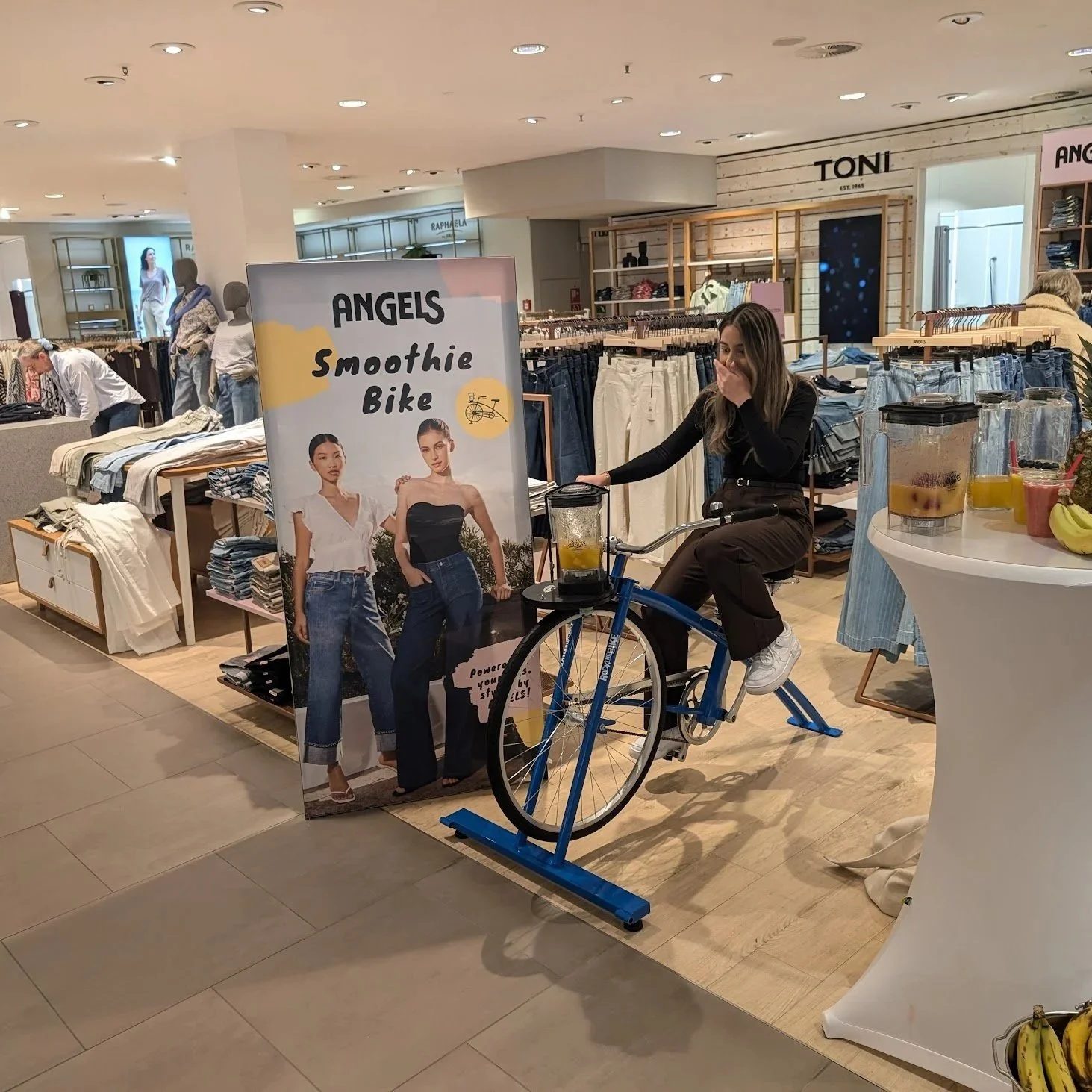 Junge Frau befindet sich auf einem stationären Fahrrad in einem Modegeschäft, daneben ein Plakat mit zwei Frauen in Jeans und T-Shirts. Es gibt mehrere Schilder und Regale mit Kleidung im Hintergrund.