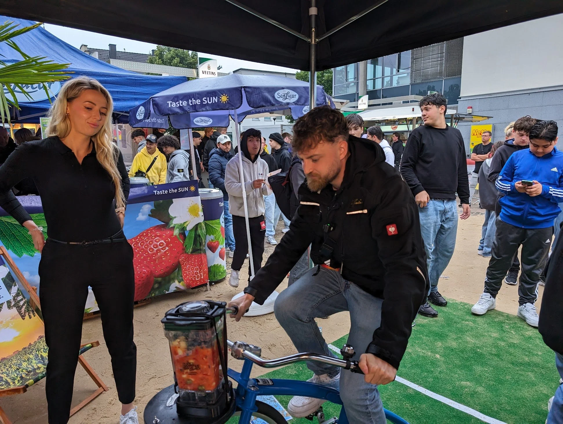 Ein Mann fährt Fahrrad auf einem Marktstand mit einem plakat, das Erdbeeren und Blumen zeigt. Eine Frau beobachtet lächelnd. Es gibt mehrere Menschen im Hintergrund, einige schauen auf ihre Handys, während andere sich umsehen.