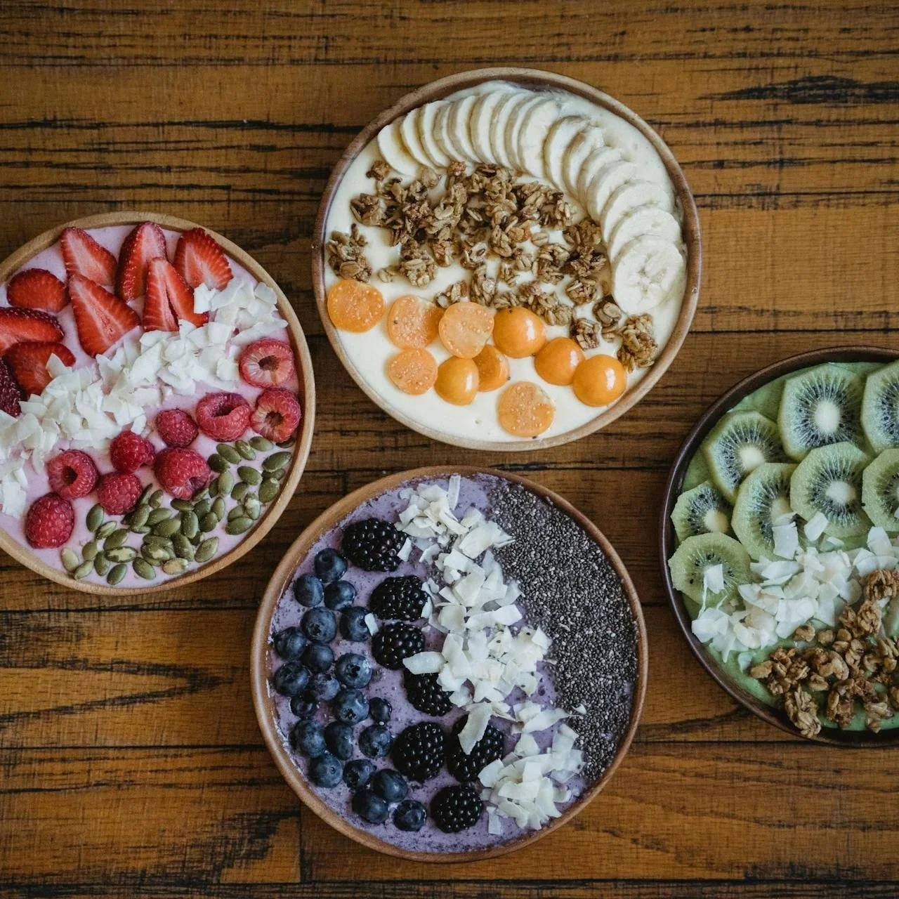Vier Schalen mit Früchte- und Beeren-Bowls, auf einem Holztisch.