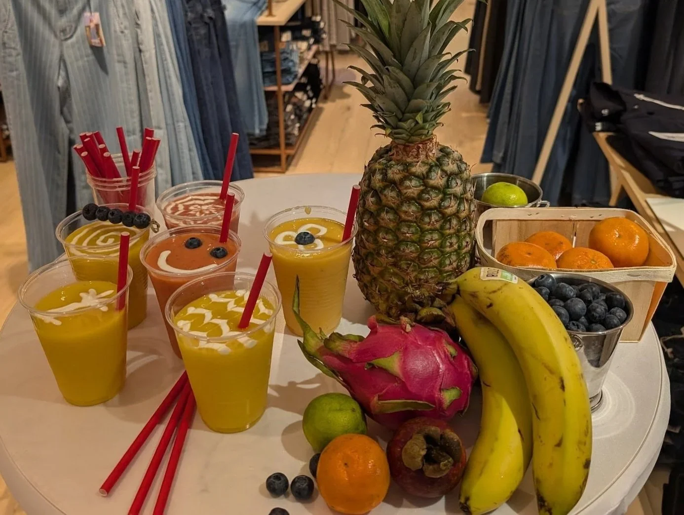 Verschiedene Gläser mit bunten Frucht-Smoothies, eine Ananas, Bananen, eine Drachenfrucht, Orangen, Limetten, Joghurt, Blaubeeren und ein Pappkorb mit weiteren Früchten auf einem Tisch.
