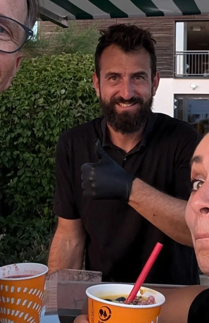 Ein lachender Mann mit dunklem Haar und Bart, trägt schwarze Kleidung und eine schwarze Handschuh, sitzt im Freien bei einem Abendessen mit anderen Personen.