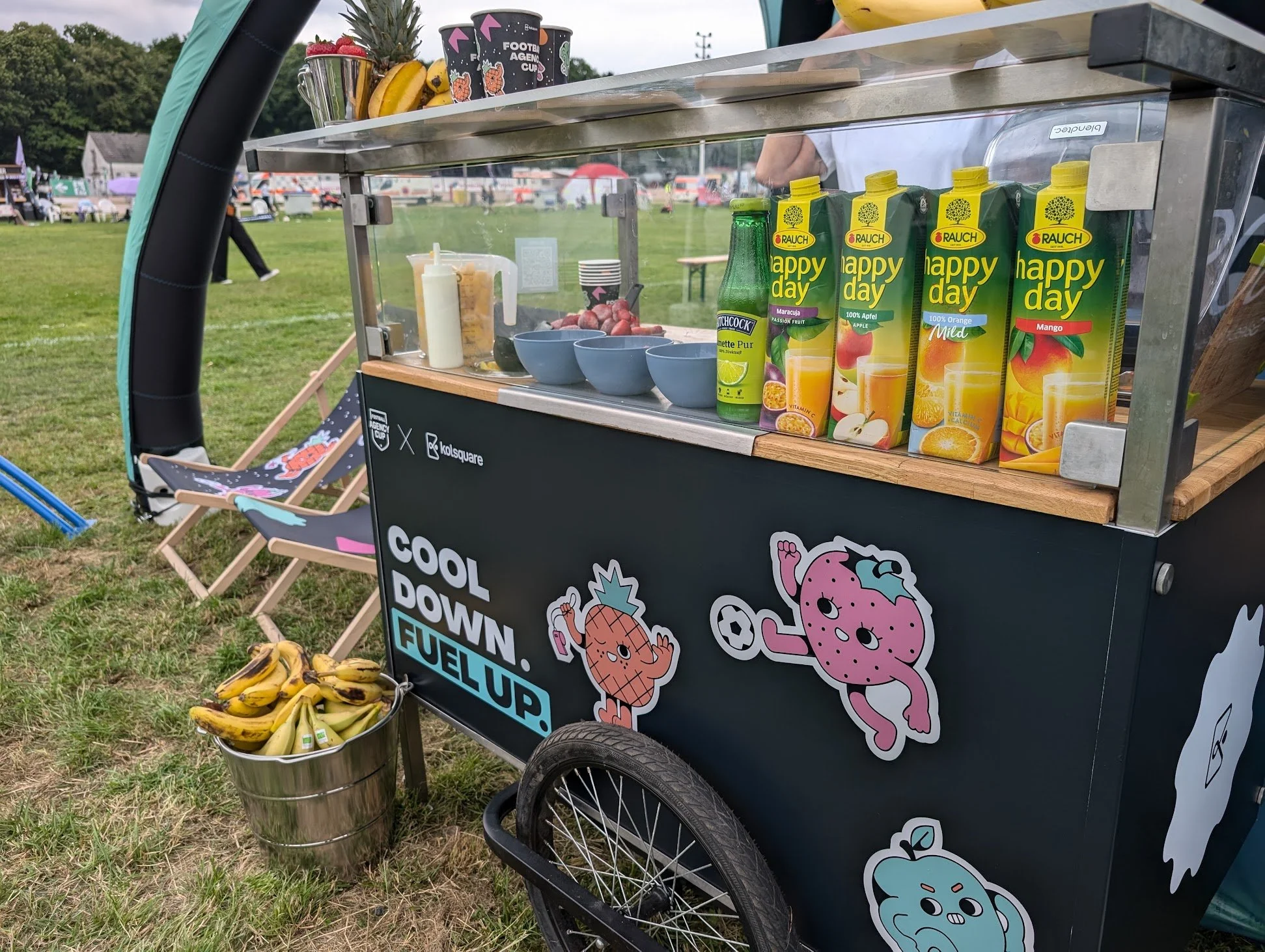 Ein Getränke- und Snackwagen im Freien mit verschiedenen Getränken, Früchten und Stickern, auf einer Wiese mit Zelten und Freizeitaktivitäten im Hintergrund.