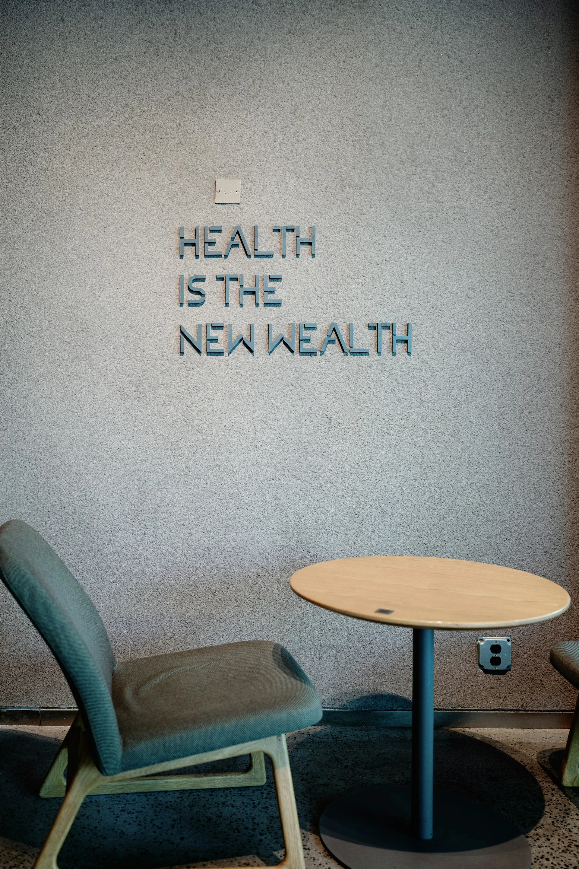 Ein grauer Sitzstuhl und ein runder Holztisch vor einer Wand mit einem Zitat auf Englisch: 'Health is the new wealth'.