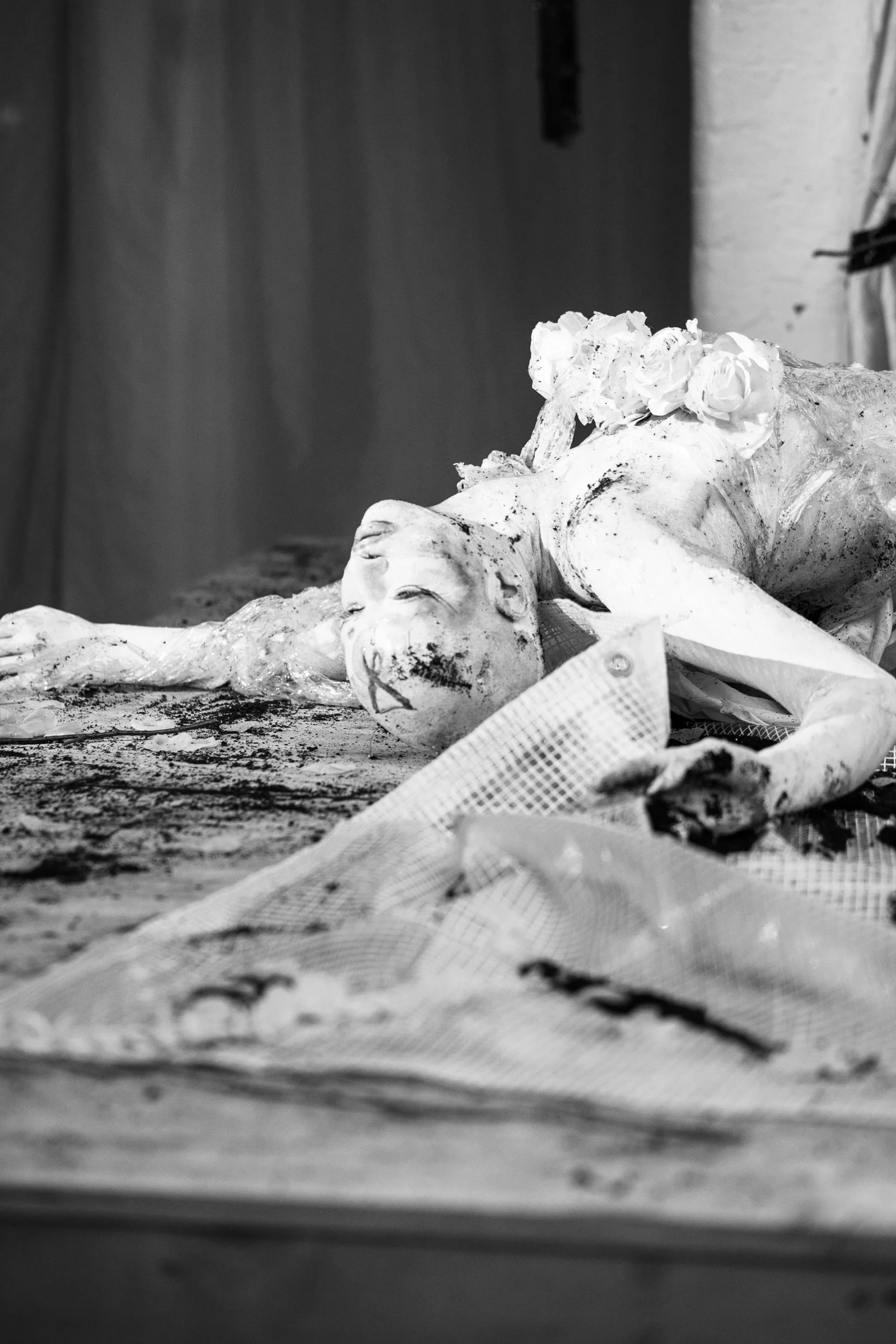 A black and white photograph of a damaged angel or saint statue lying on a table or altar, with a wreath of flowers on its head.