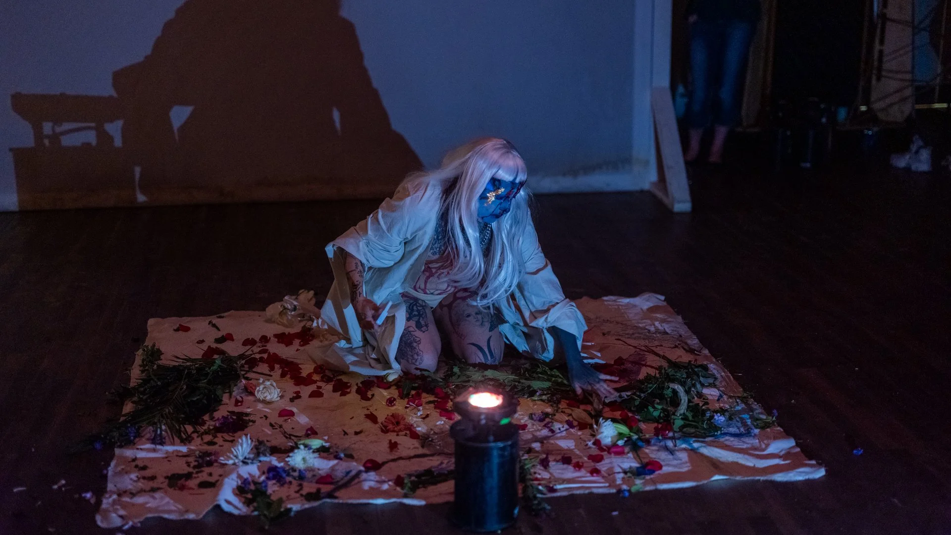 A person with long blonde hair kneeling on a cloth with flower petals and greenery, wearing a light-colored coat and a face mask, illuminated by a candle, in a dimly lit room.