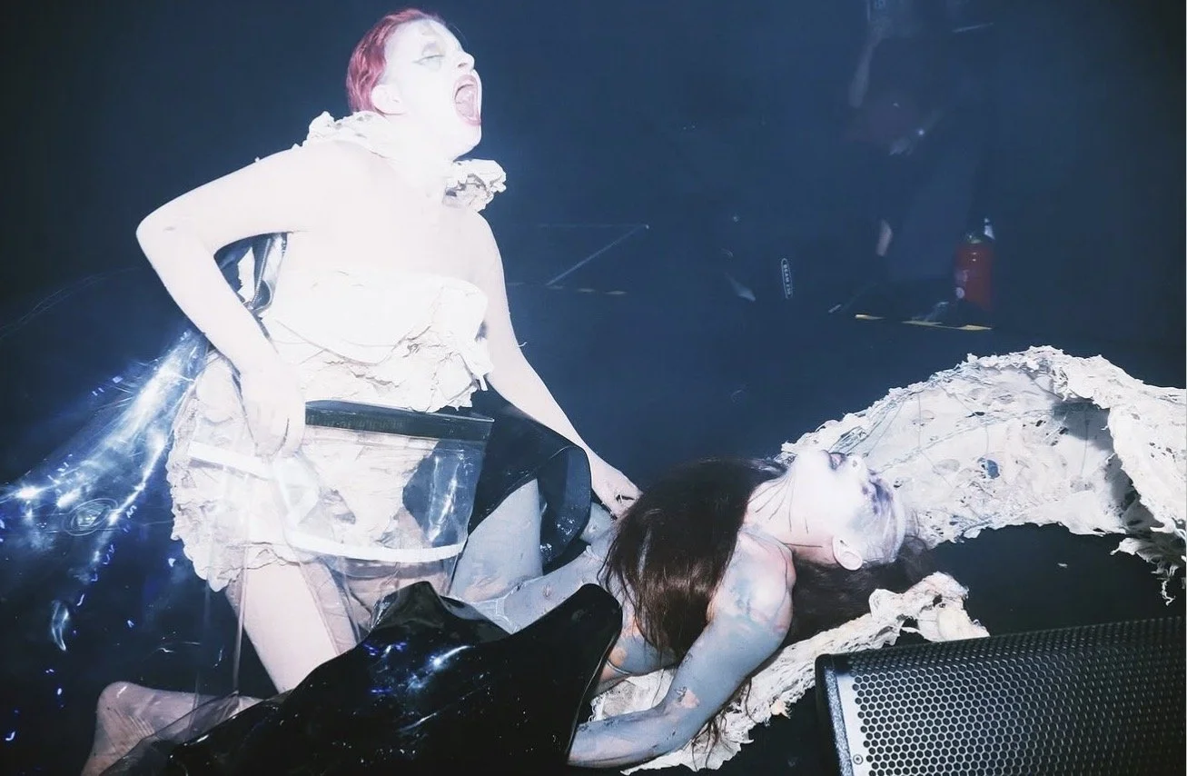 Art installation resembling two women in a dark space, one with red hair screaming and the other lying down, with torn paper-like material around them.