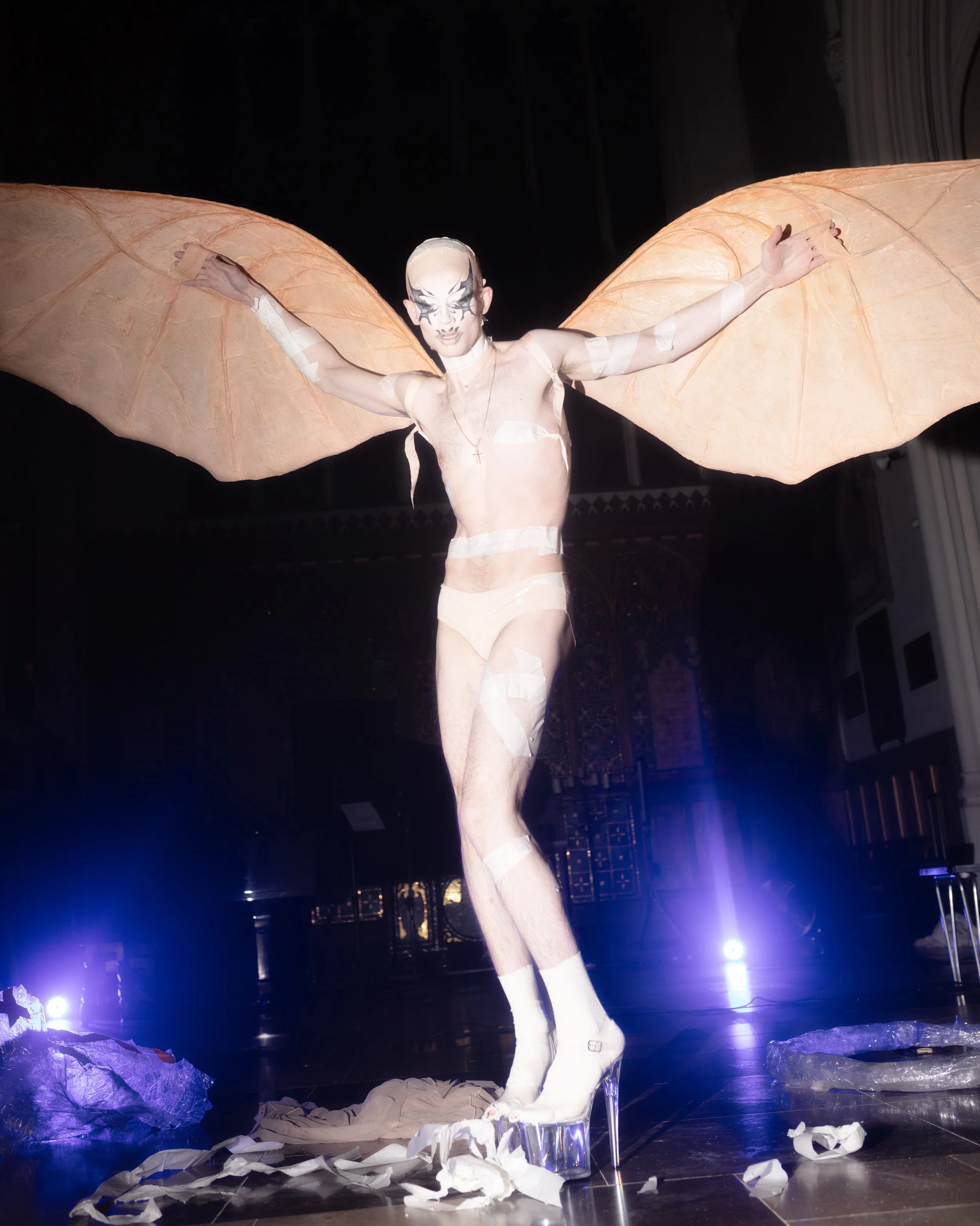 A person with elaborate makeup and large orange wings stands on stage with arms outstretched, wearing sheer clothing, high heels, and surrounded by scattered paper.