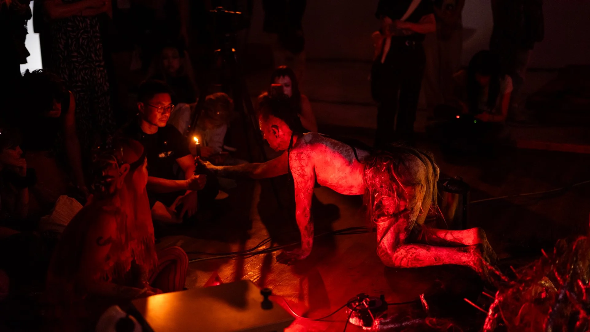A performer dressed as a mythical creature with a naked upper body and elaborate costume, crawling on all fours on a dark stage illuminated by red light, with an audience sitting and watching. The performer has long black hair and painted skin, creat