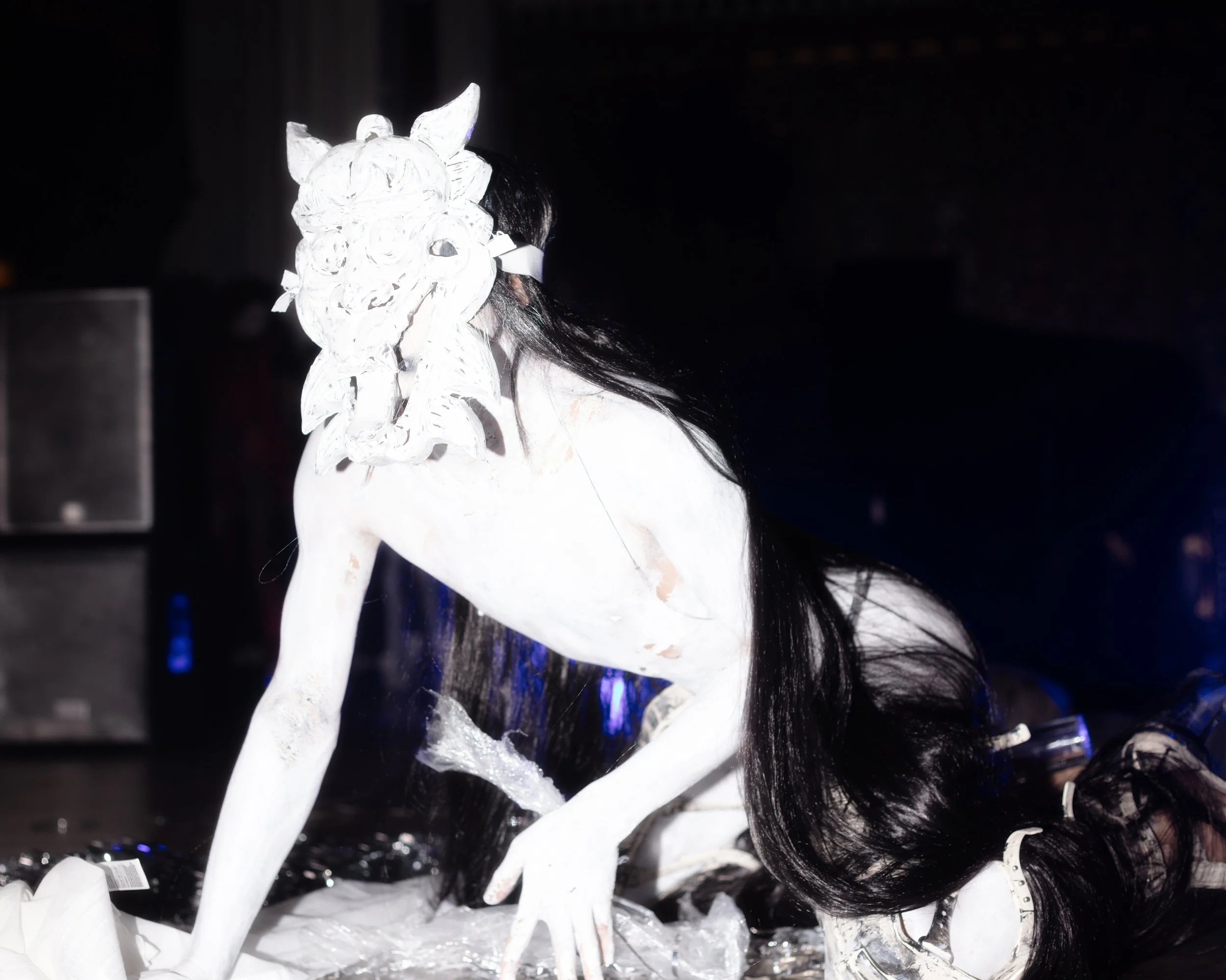 A person wearing a mask resembling a lion's face, with a mane, crouching on the floor in a dark setting.