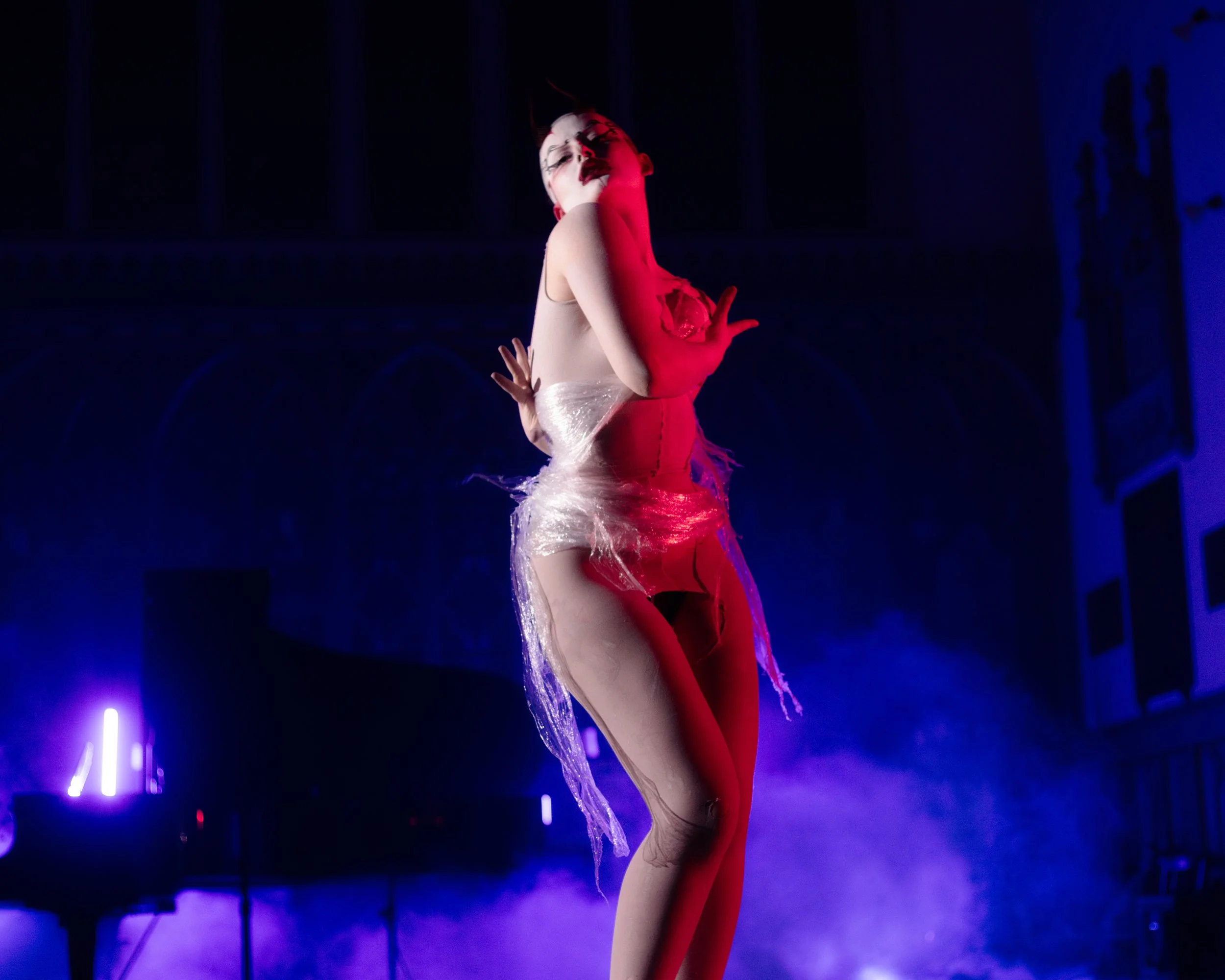 Performer in a dramatic pose on stage, lit with colorful stage lights and fog.
