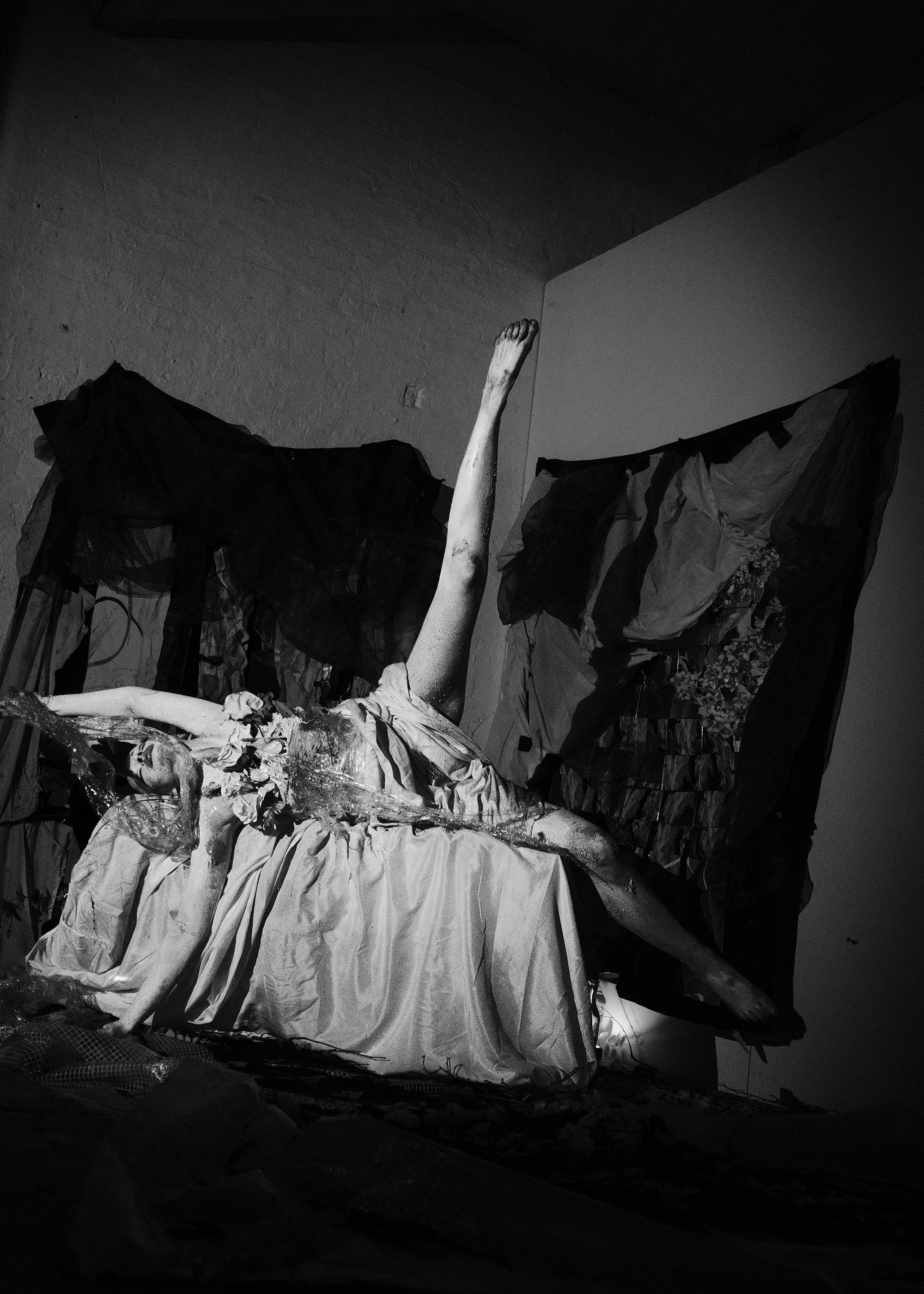 Black and white photograph of an abstract sculpture of a reclining woman with one arm raised. The sculpture appears to be made of mixed materials, with fabric and textured surfaces, placed against a textured wall with fabric or paper drapes in the ba