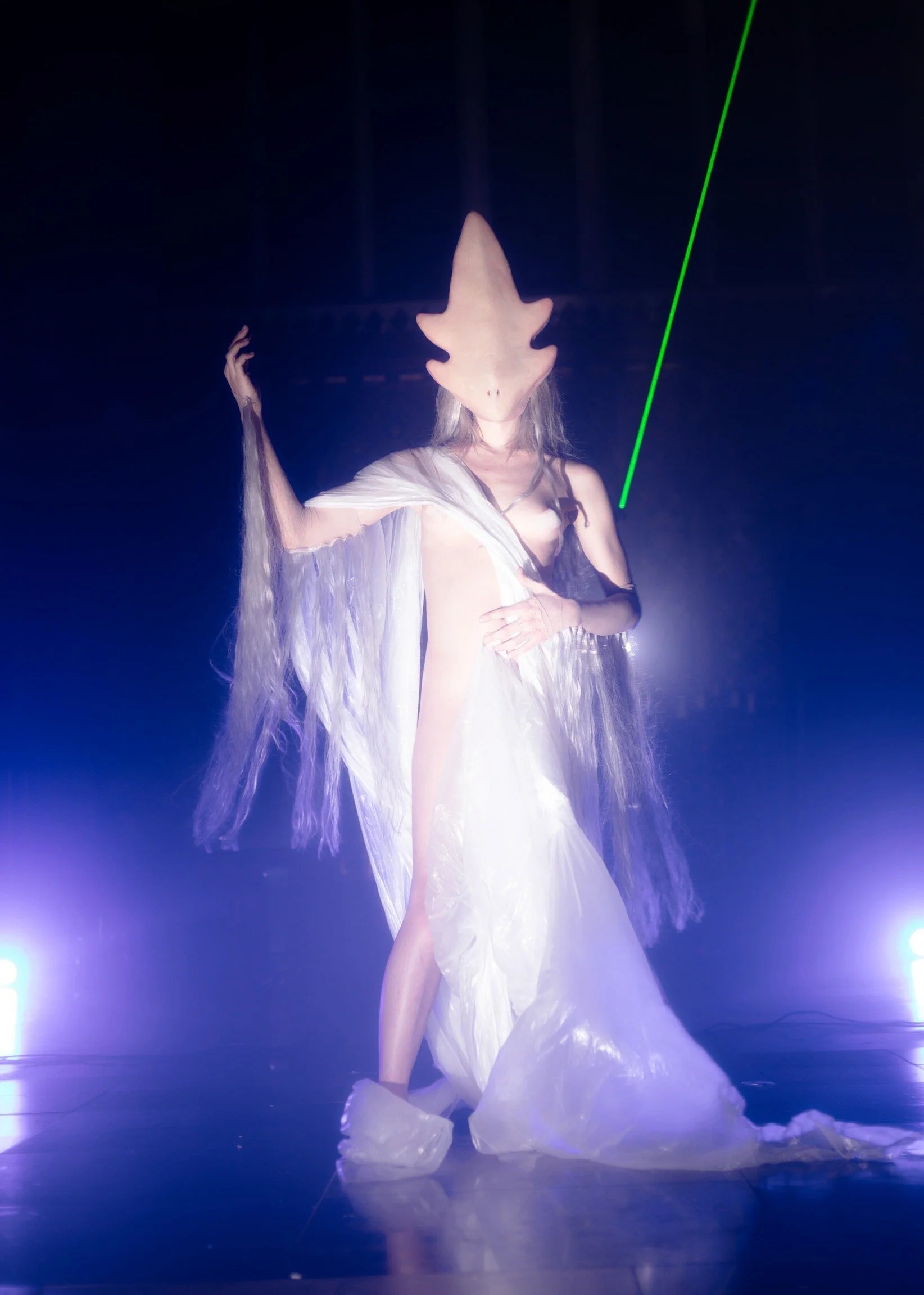 A person wearing a large, shark-shaped mask and a flowing white dress with long, wavy fabric extensions, standing on stage with blue and purple lights. The person has one hand on their chest and the other raised, amid a misty or foggy atmosphere.