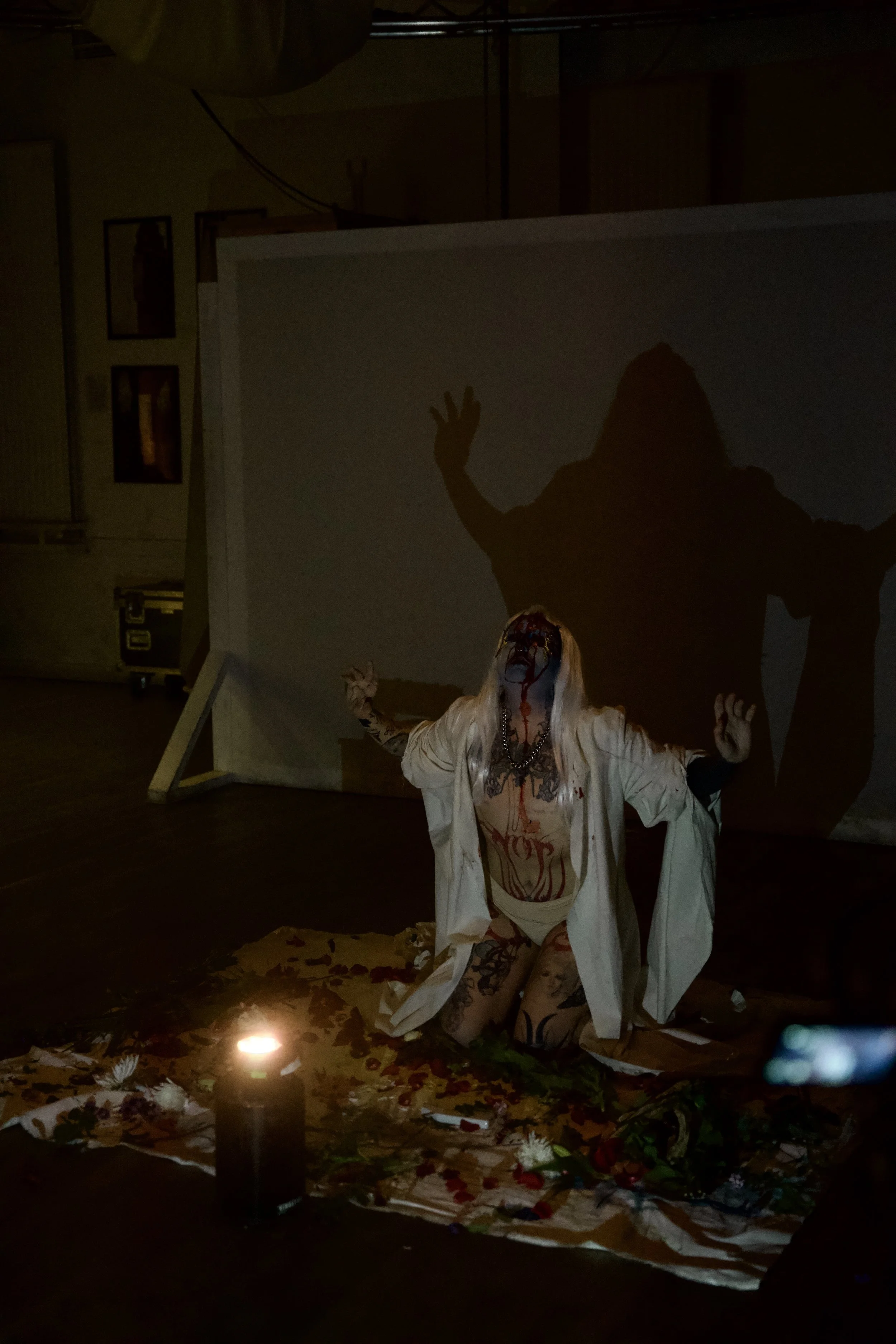 A person with long blonde hair and tattoos, dressed in white, kneeling on a cloth with flowers and leaves, illuminated by a small candle, with their face covered in fake blood, in front of a large shadow cast on a backdrop.