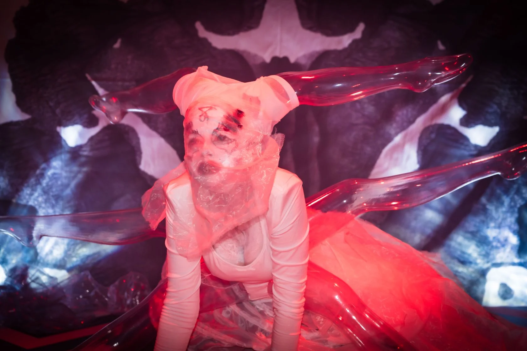 Person with face tattoos wearing a sheer pink top and plastic head covering, surrounded by red and clear plastic sculptures, with dark abstract background.
