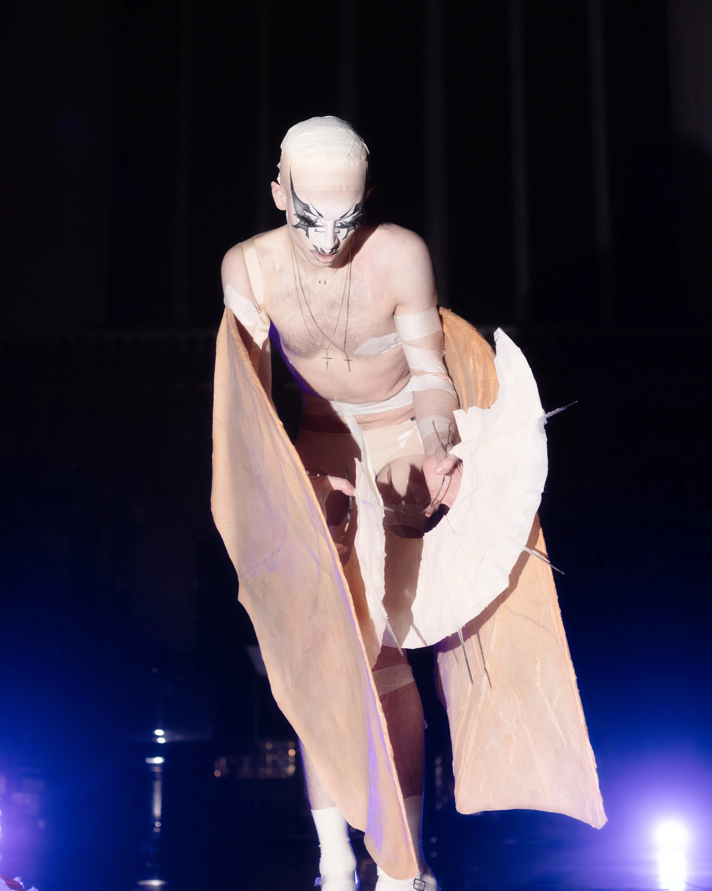 A performer with pale skin, wearing a white mask with black designs, and a cross necklace, is on stage in a minimal costume with large, light-colored wings or fabric attached to their arms, illuminated by stage lights.