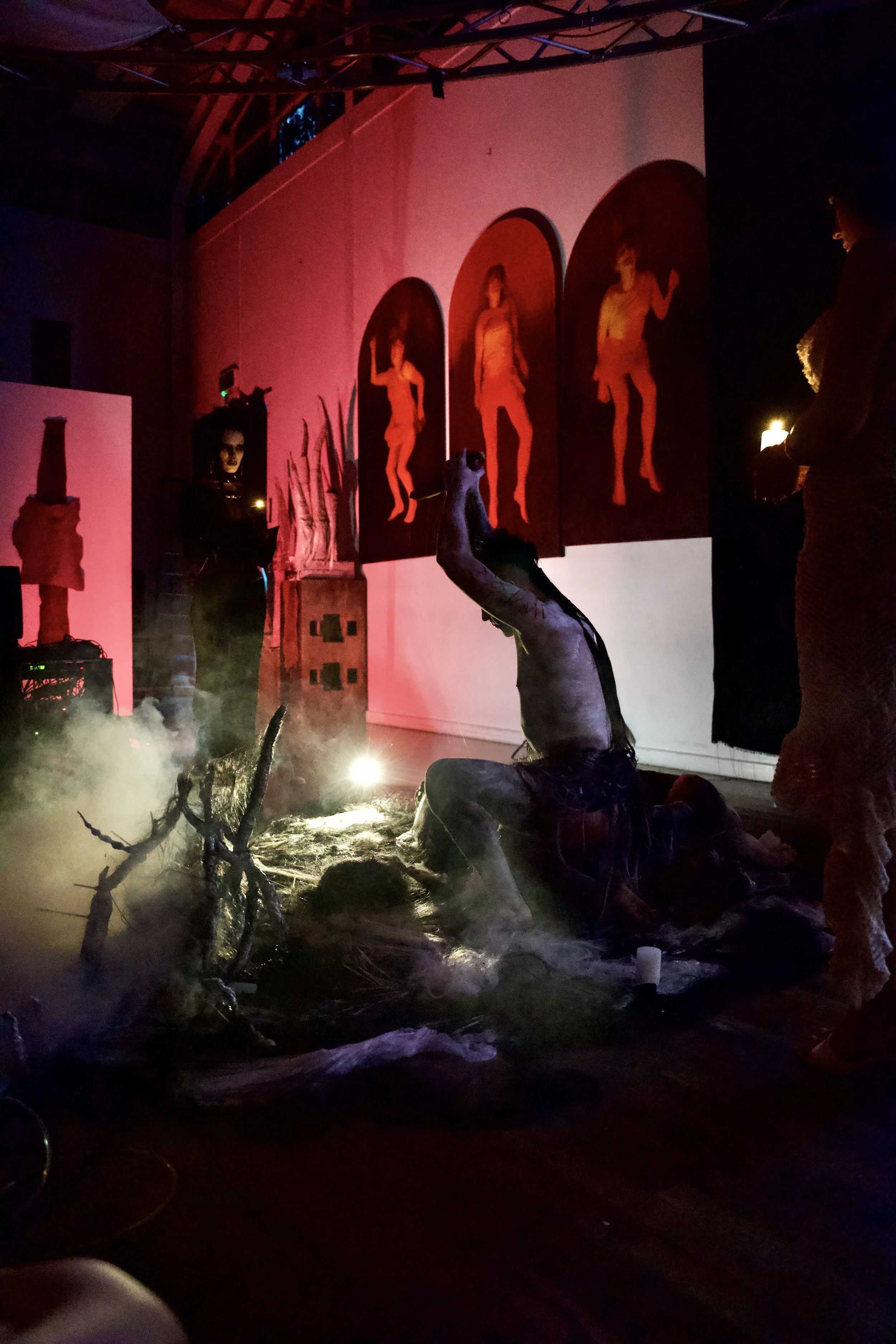 A dimly lit room with horror-themed decorations including a person in a creepy costume with a large, distorted head, smoke effects, and eerie lighting. There are four red-clad figures on the wall, with a few people standing nearby.