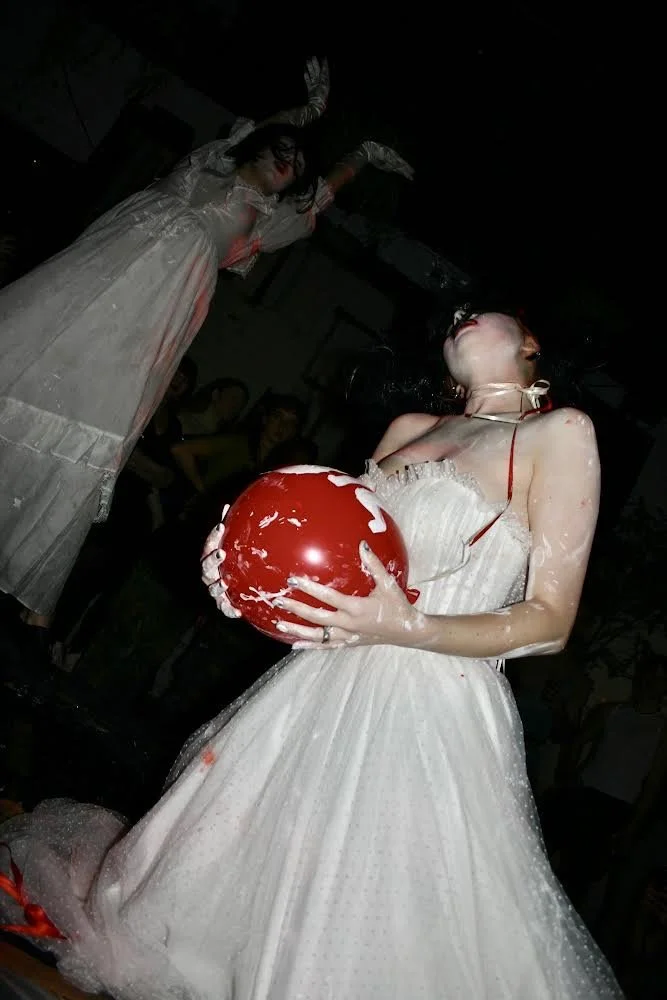 A woman in a white dress holding a red balloon with white foam on it, with another woman in the background wearing a white dress and white gloves, in a dark room.