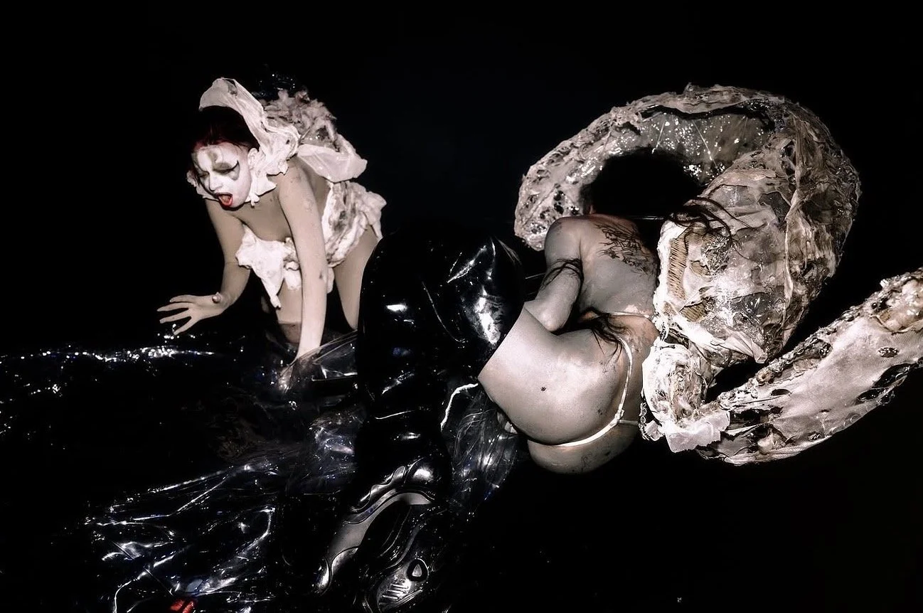 Two people dressed in avant-garde costumes with mixed textures and materials, posing on a black background.