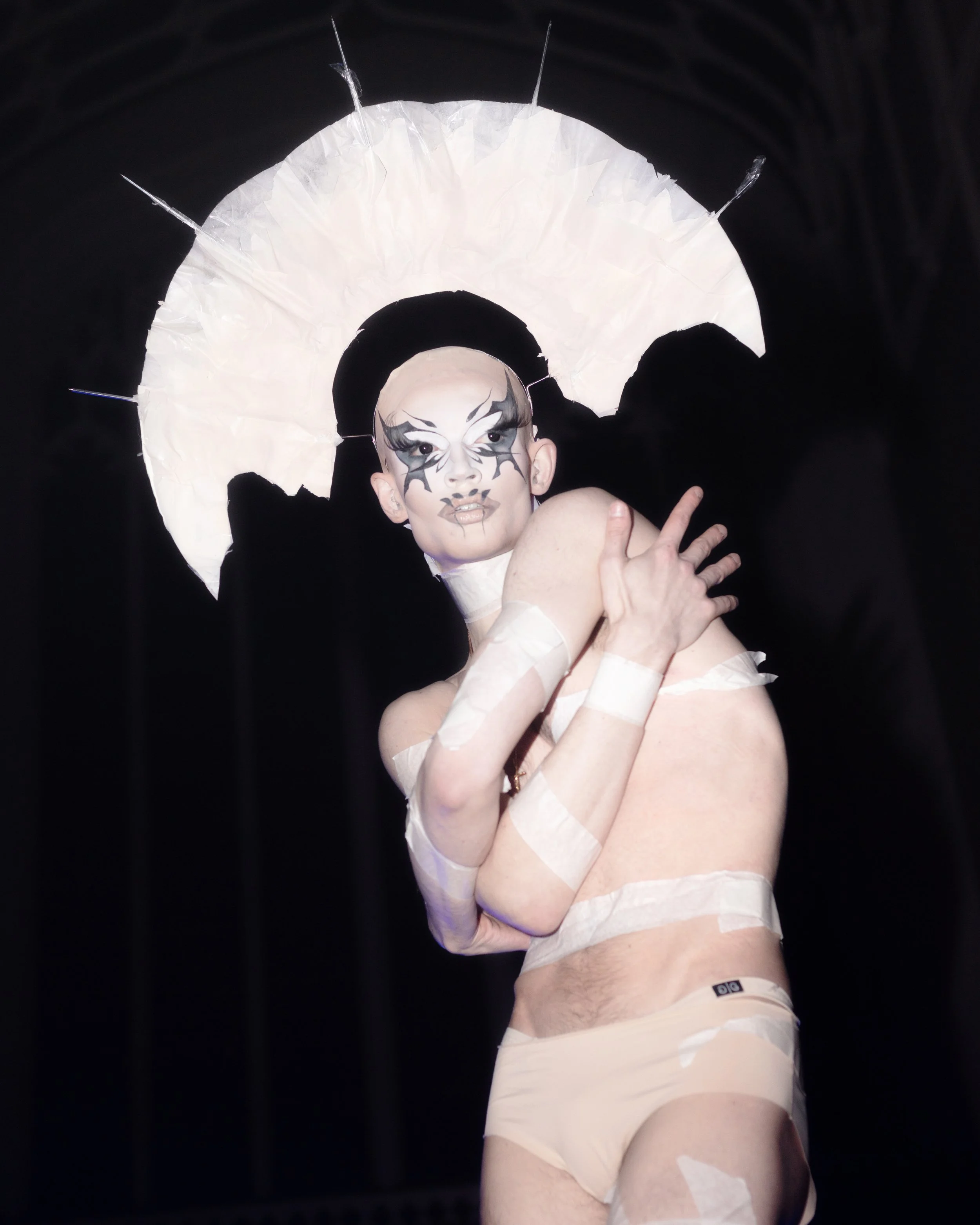 Person dressed in elaborate makeup, wearing a headpiece with an umbrella-like structure, pose with arms crossed over shoulders during an avant garde performance live show in London. 